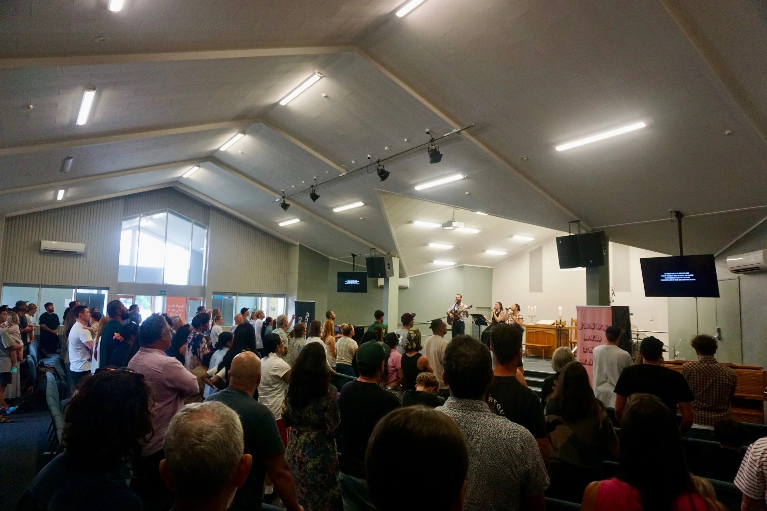 A diverse group of people gathered in a church or event hall, watching a band perform on stage with two singers and a guitarist, while screens display lyrics or messages.