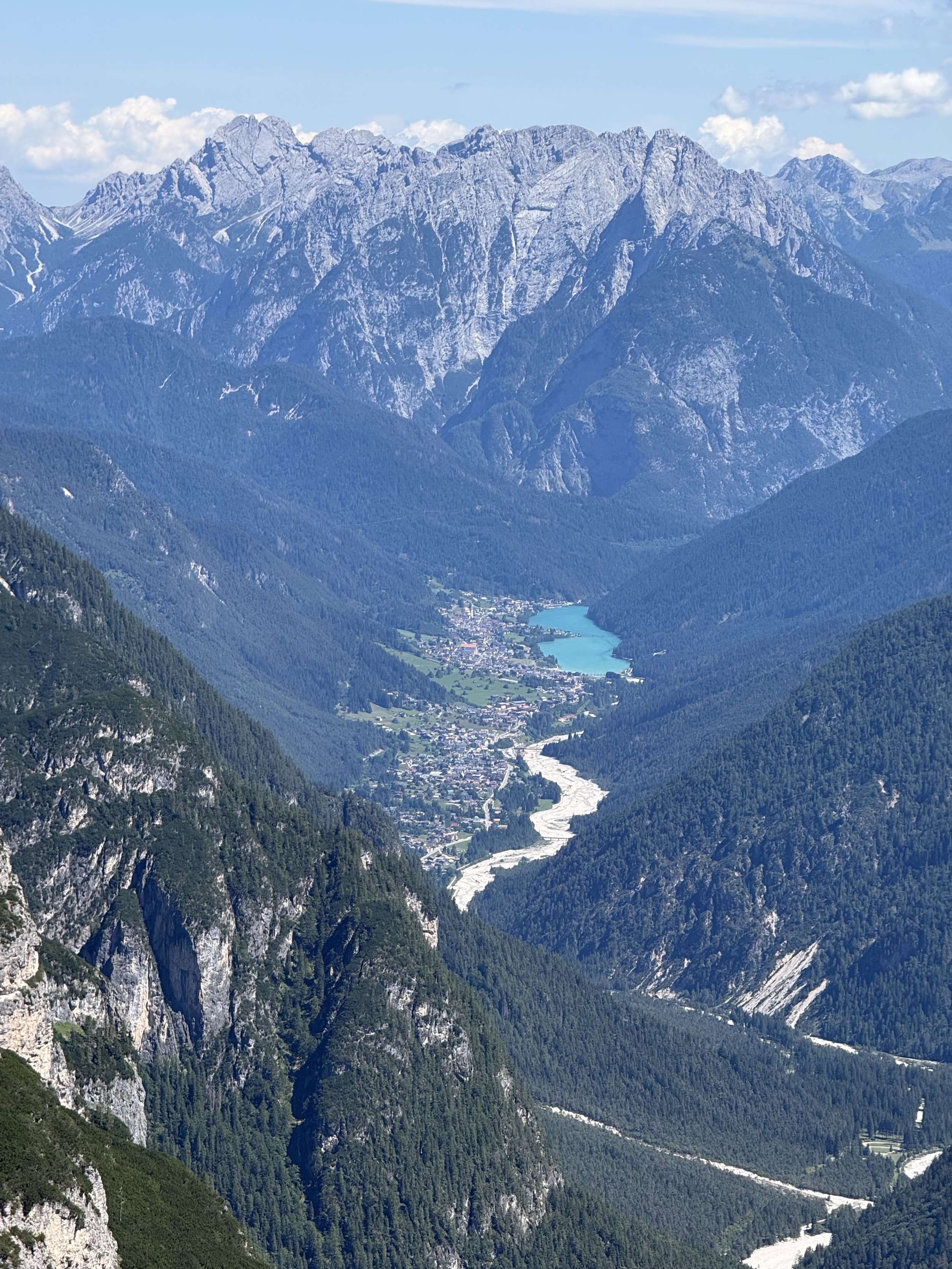 A panoramic view of a mountainous landscape with tall, rugged peaks and a small village nestled in a valley near a river, surrounded by lush green forests.