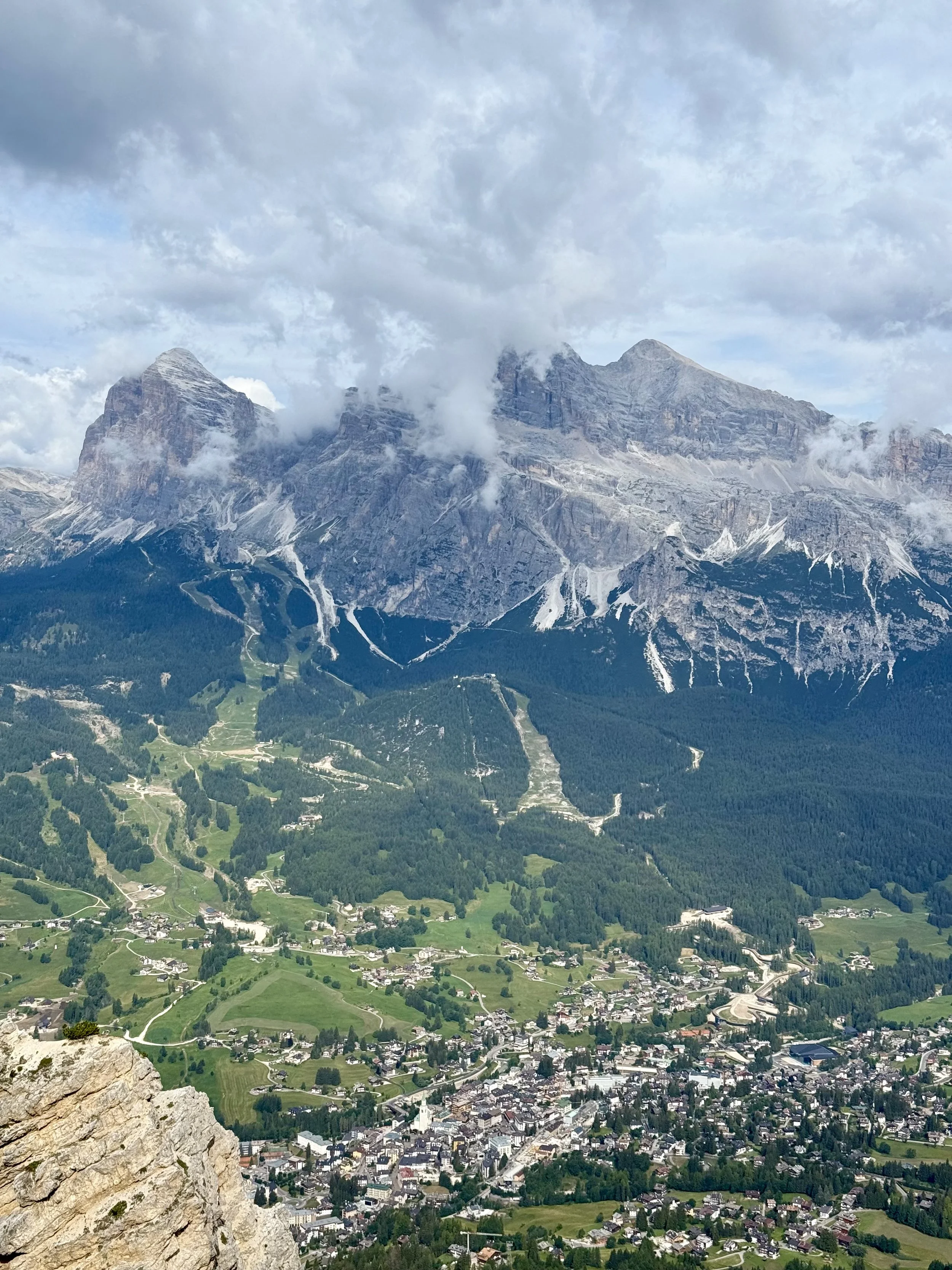 A mountainous landscape with rugged peaks partially covered in clouds and snow, overlooking a green valley with a small town and winding roads.