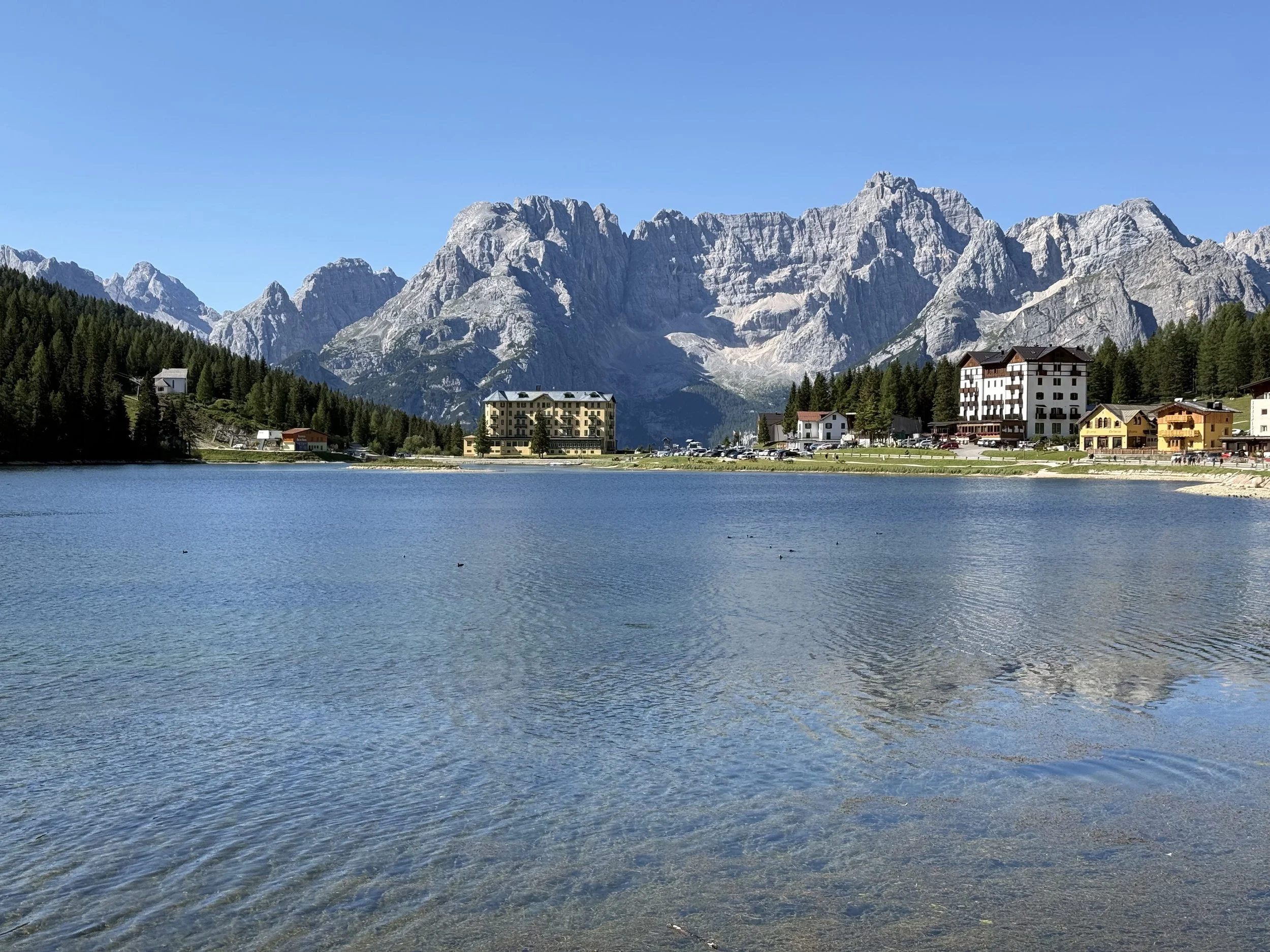 A serene mountain village with colorful buildings near a calm lake, surrounded by dense green forest and towering gray rocky mountains under a clear blue sky.