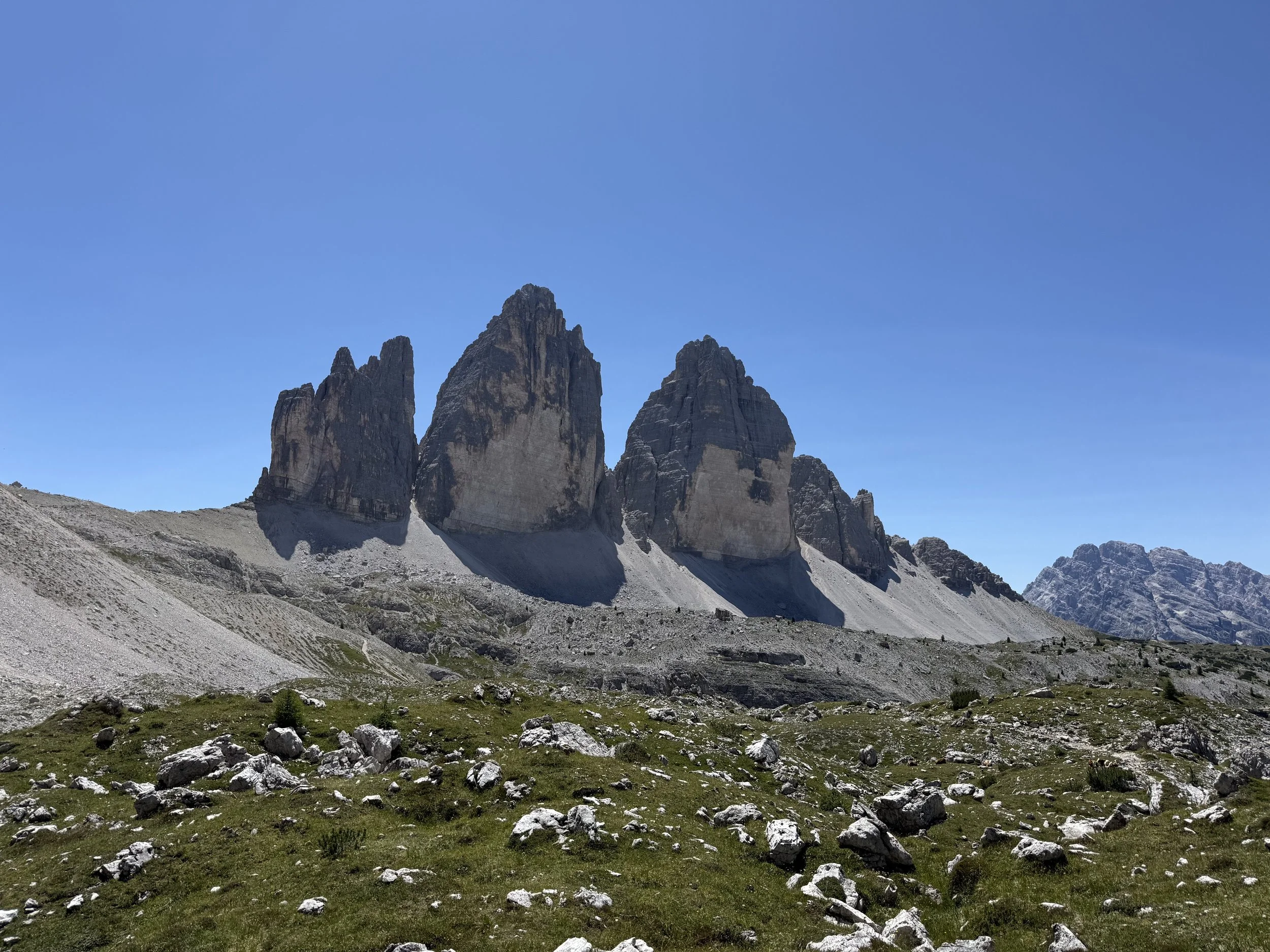 A mountain range with three prominent peaks under a clear blue sky, rocky terrain, and sparse vegetation in the foreground.