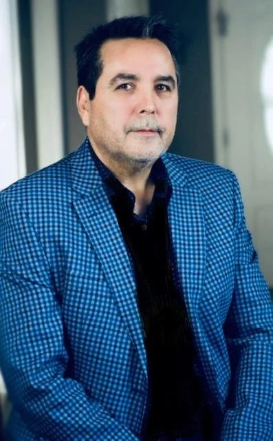 Man with dark hair and beard wearing a blue checkered blazer and black shirt, standing indoors near a window or light source.