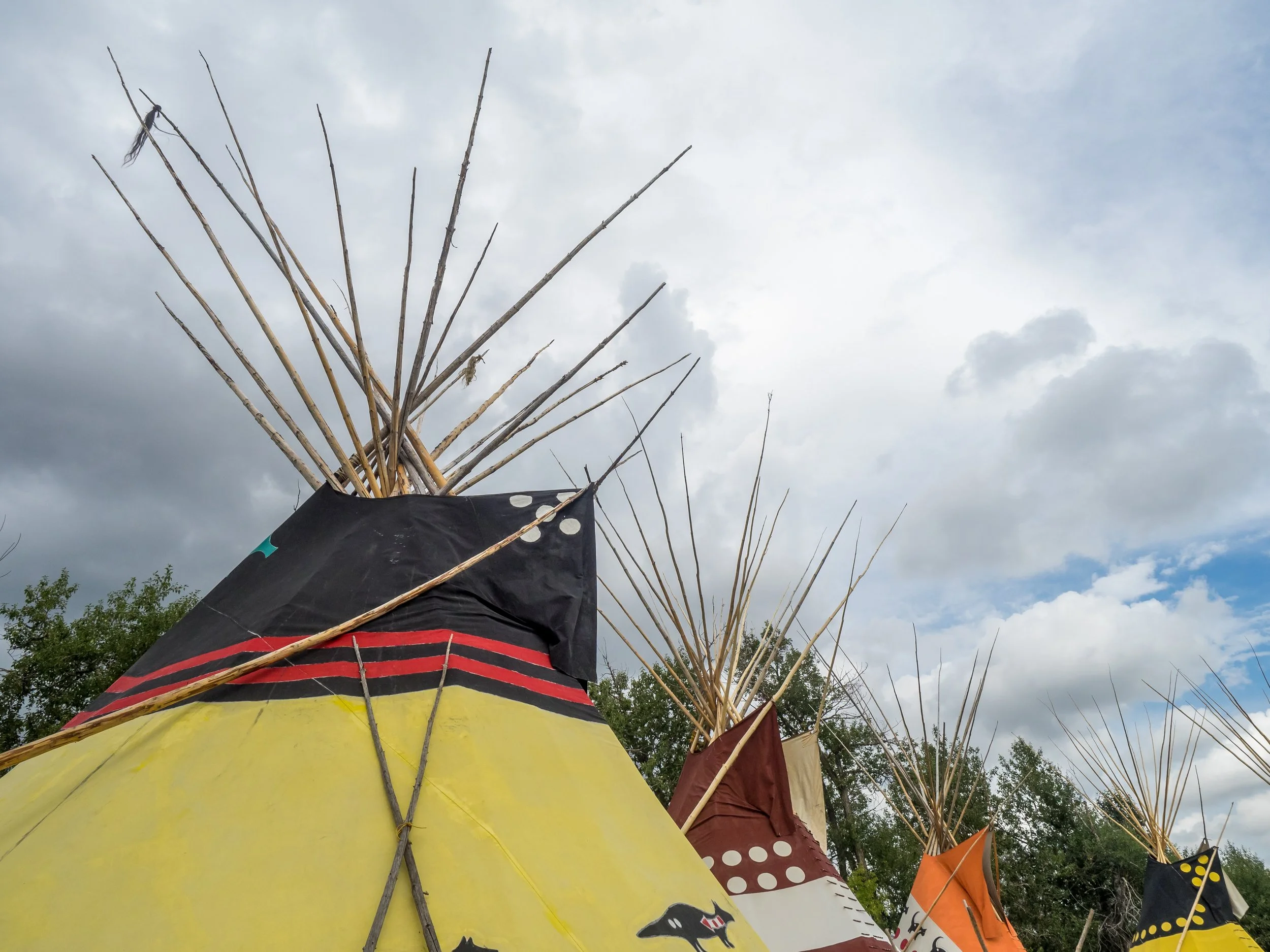 Several decorated tipis with wooden poles extending upward, set against a cloudy sky and a few trees in the background.