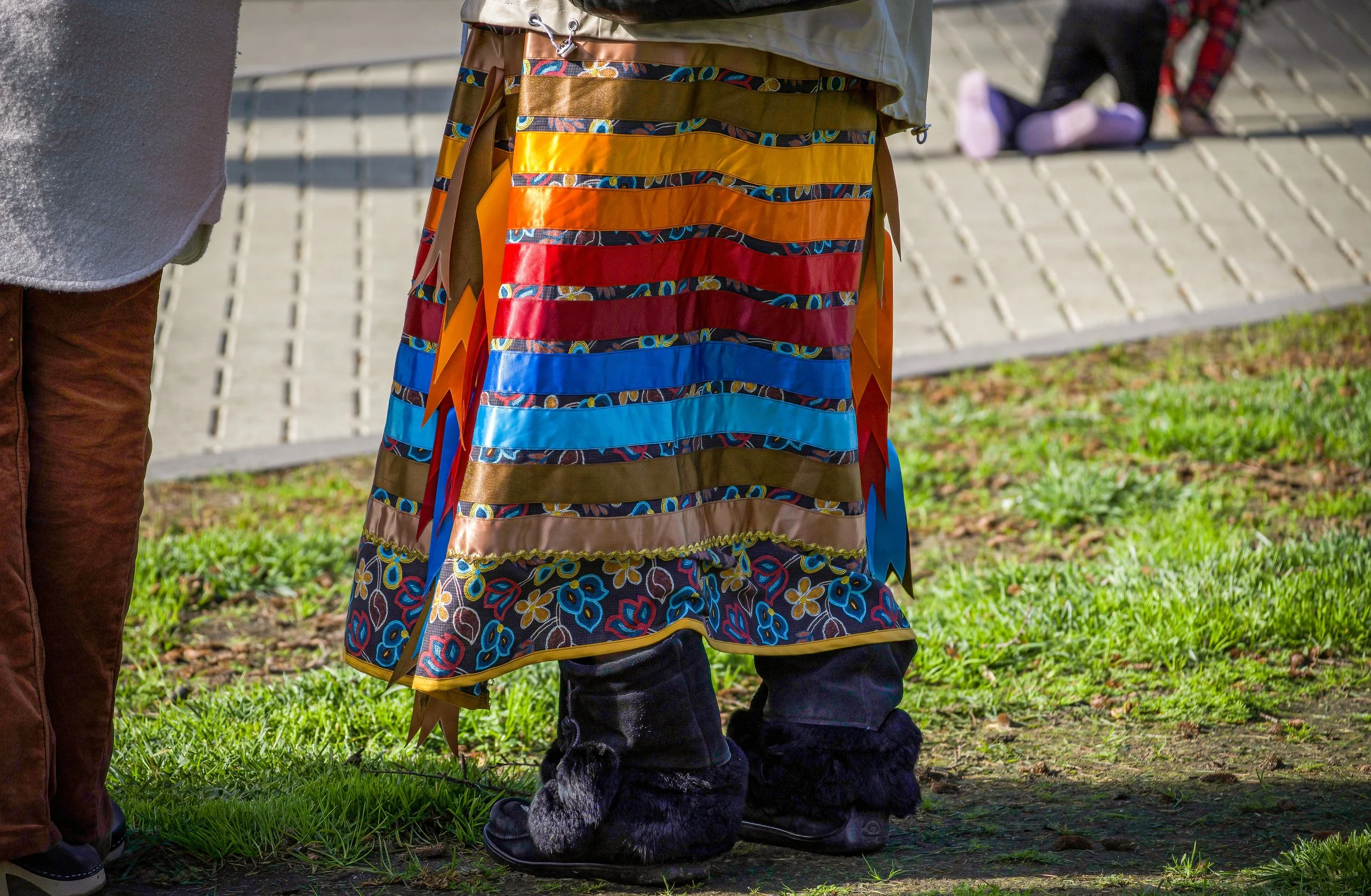 A person wearing traditional indigenous clothing with a colorful, patterned skirt and ribbon decorations stands outdoors on grass near a sidewalk.
