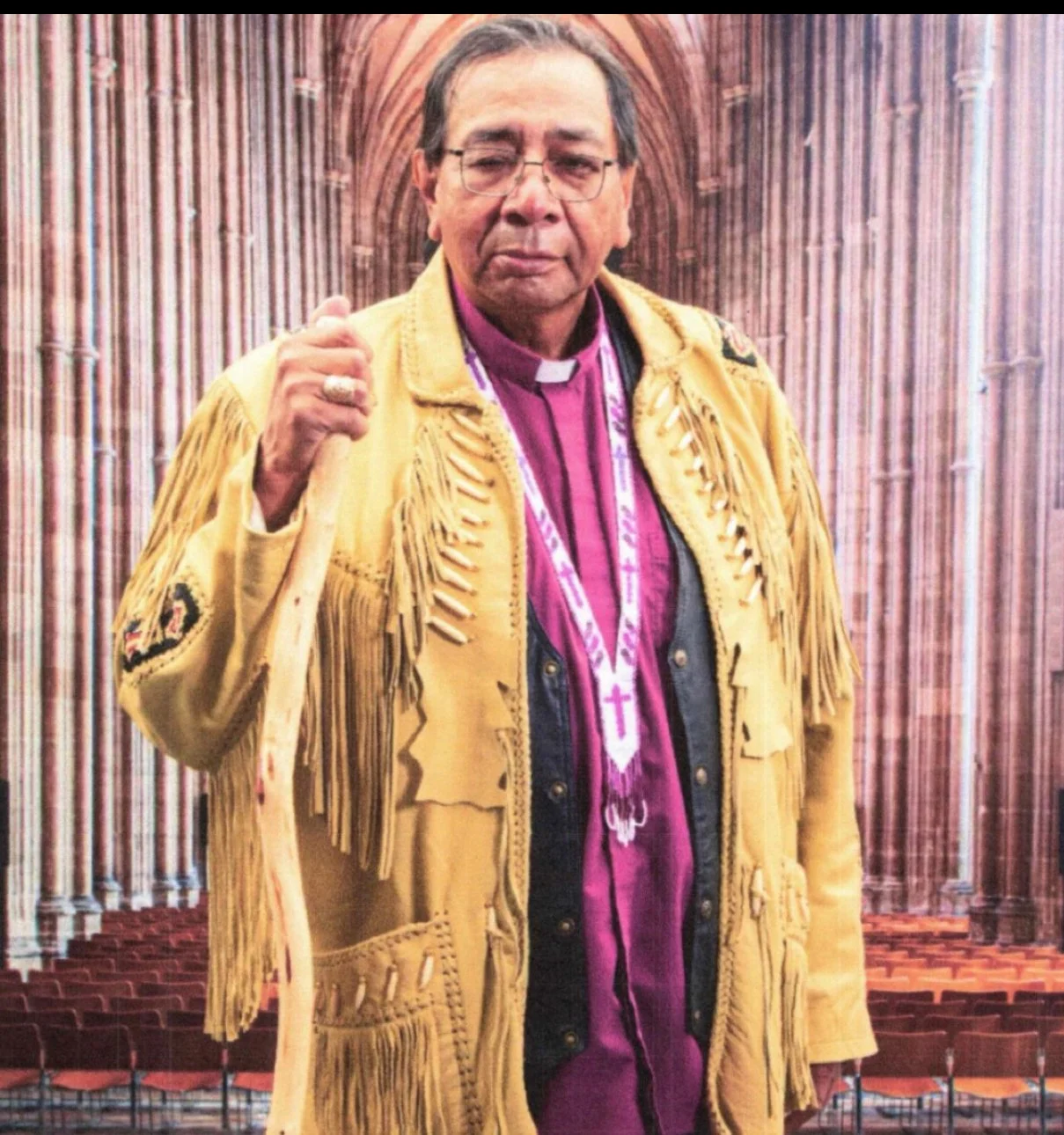 Smiling elderly man wearing a black jacket and orange shirt outdoors with green hills and a cloudy sky in the background.