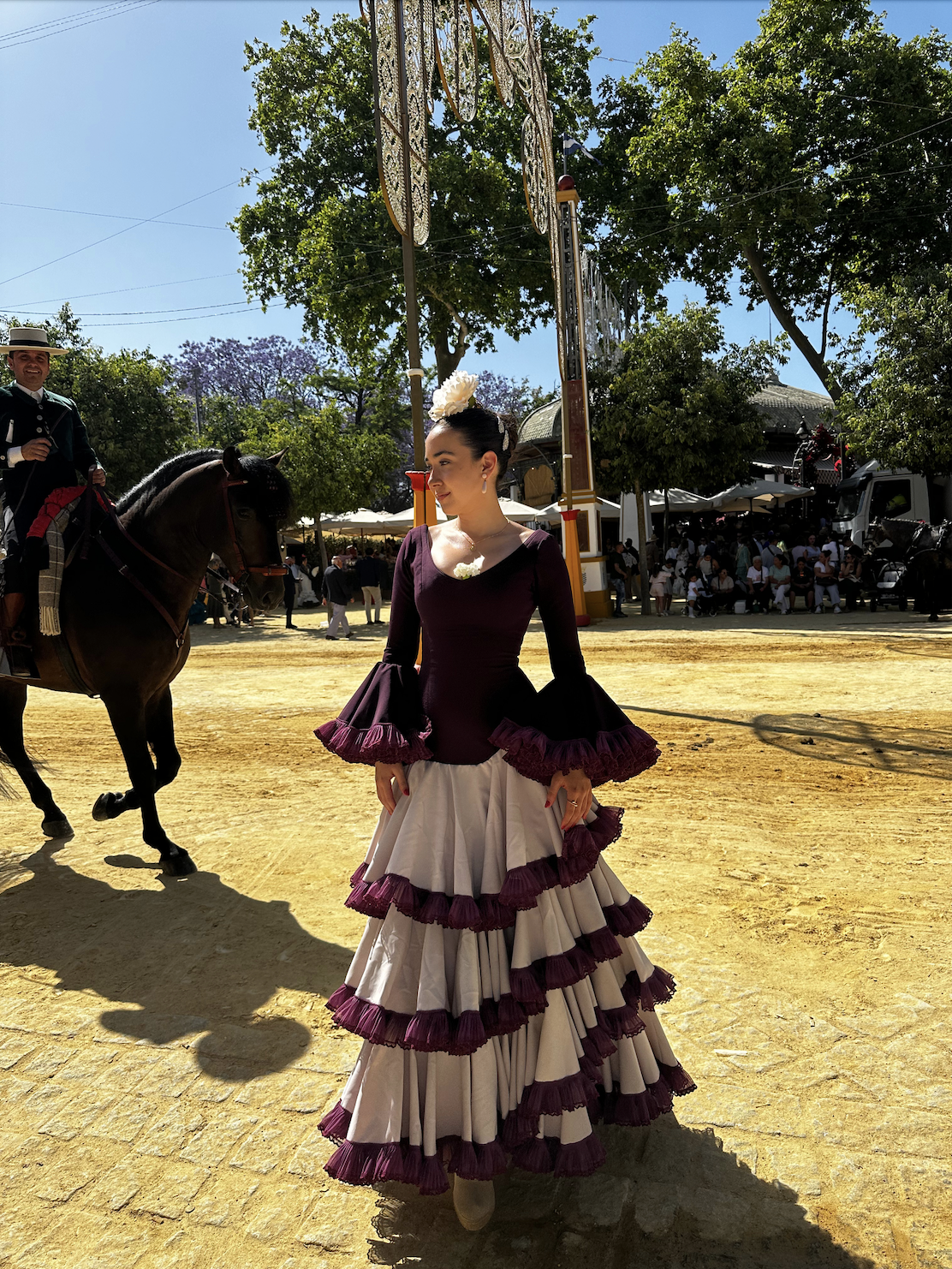A woman dressed in traditional Spanish attire with a long, ruffled dress standing outdoors during a festival or celebration. In the background, there are people, trees, and a horse with a rider, who is also wearing traditional clothing. The woman is smiling and looking to her left.