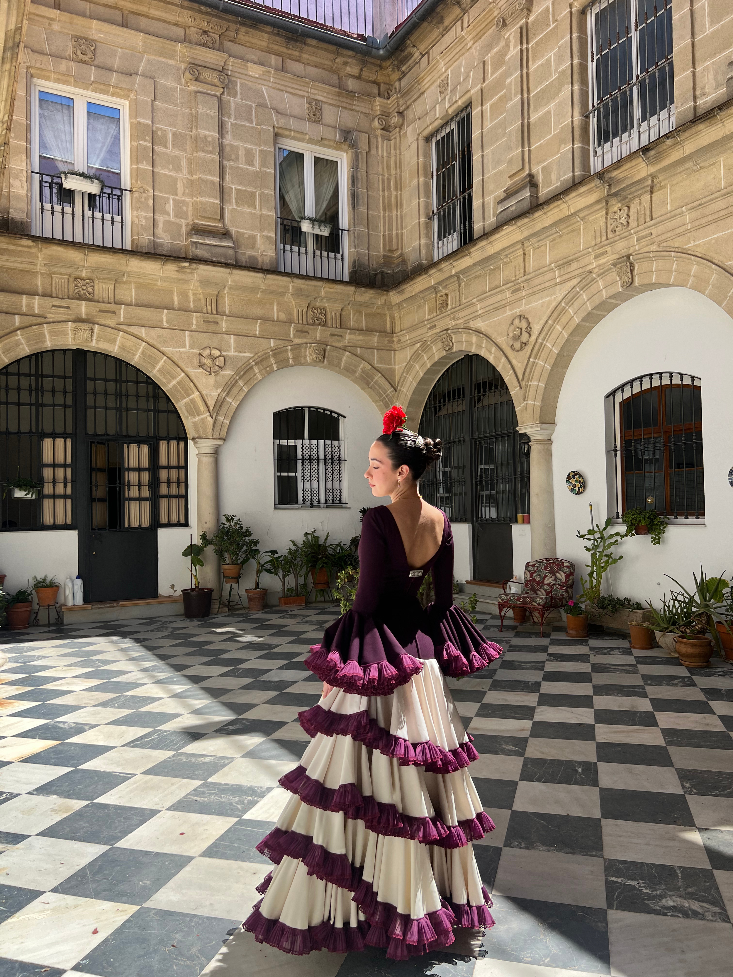 A woman in traditional Spanish dress, featuring a black fitted top with flared sleeves and a long layered skirt with purple and white ruffles, stands in a sunlit courtyard with checkered black-and-white tile flooring, potted plants, and old stone buildings with arches and barred windows.