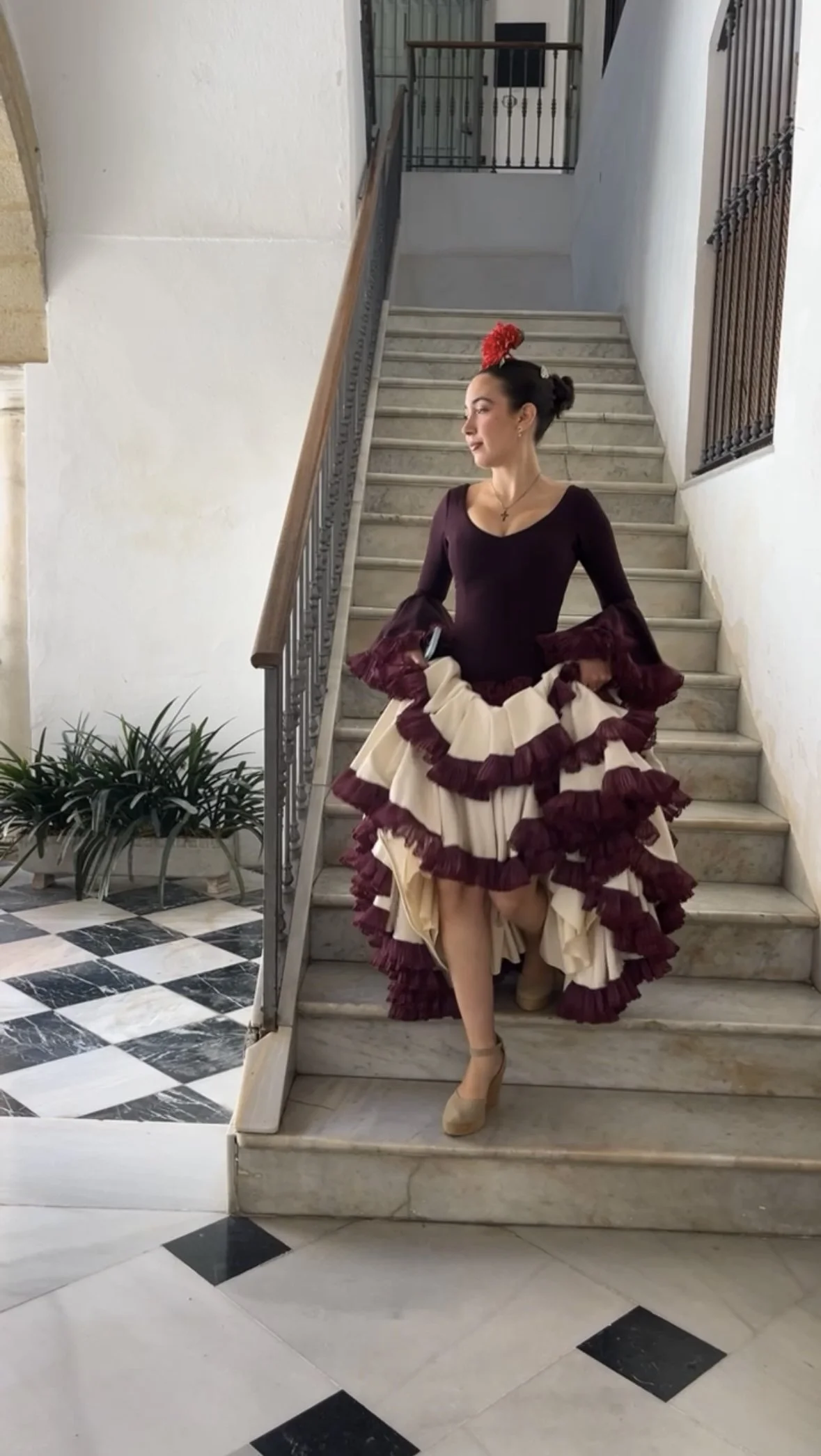 Woman in traditional flamenco dress standing on marble staircase indoors, with plants and checkered floor nearby.