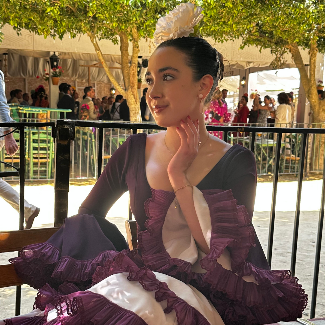 A woman in traditional Mexican dress with ruffled sleeves and a large white flower in her hair, sitting in a festive outdoor setting.