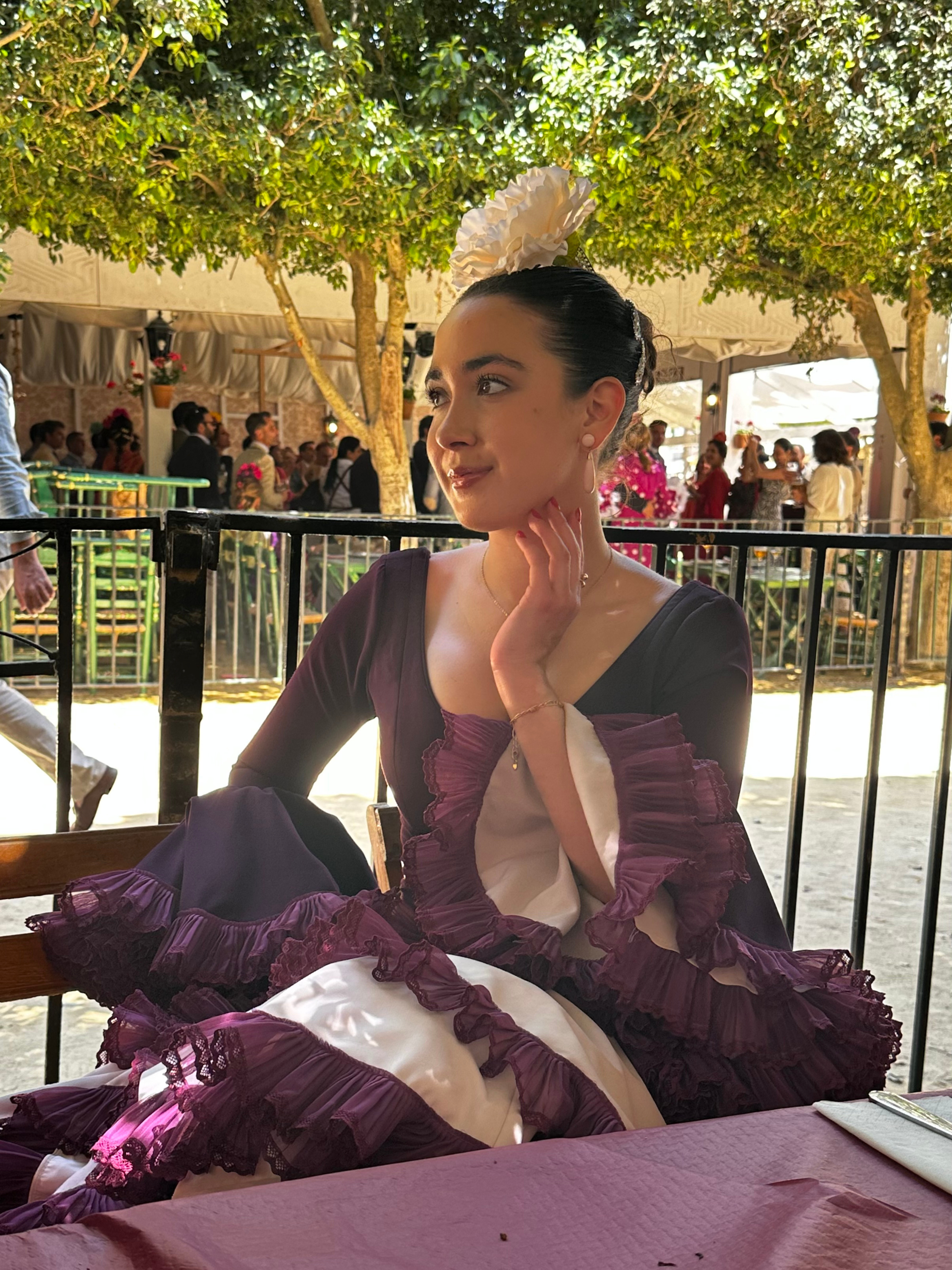 A woman dressed in traditional Mexican attire, sitting at a table with a pink tablecloth, at a festive event outdoors. She has dark hair styled in an updo with a large white floral hairpiece, and wears a black dress with purple and white ruffled accents.