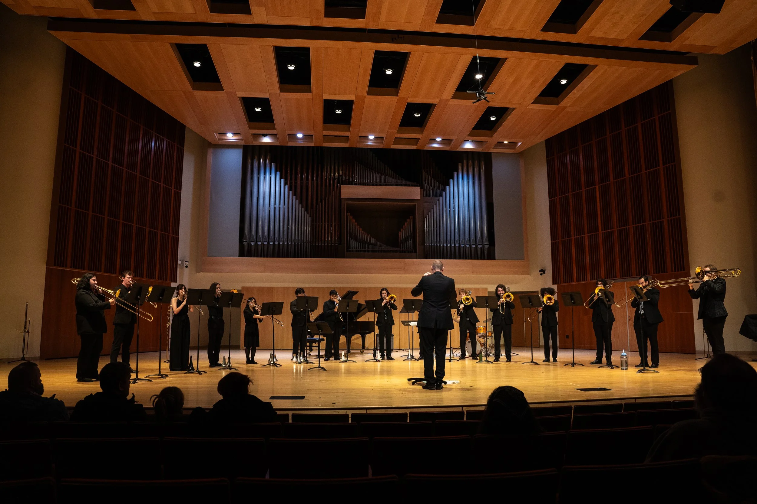The Ithaca College Trombone Troupe performing on the Ford Hall stage. Dr. Will Porter is conducting an ensemble of tenor trombones, bass trombones, and a contrabass trombone.