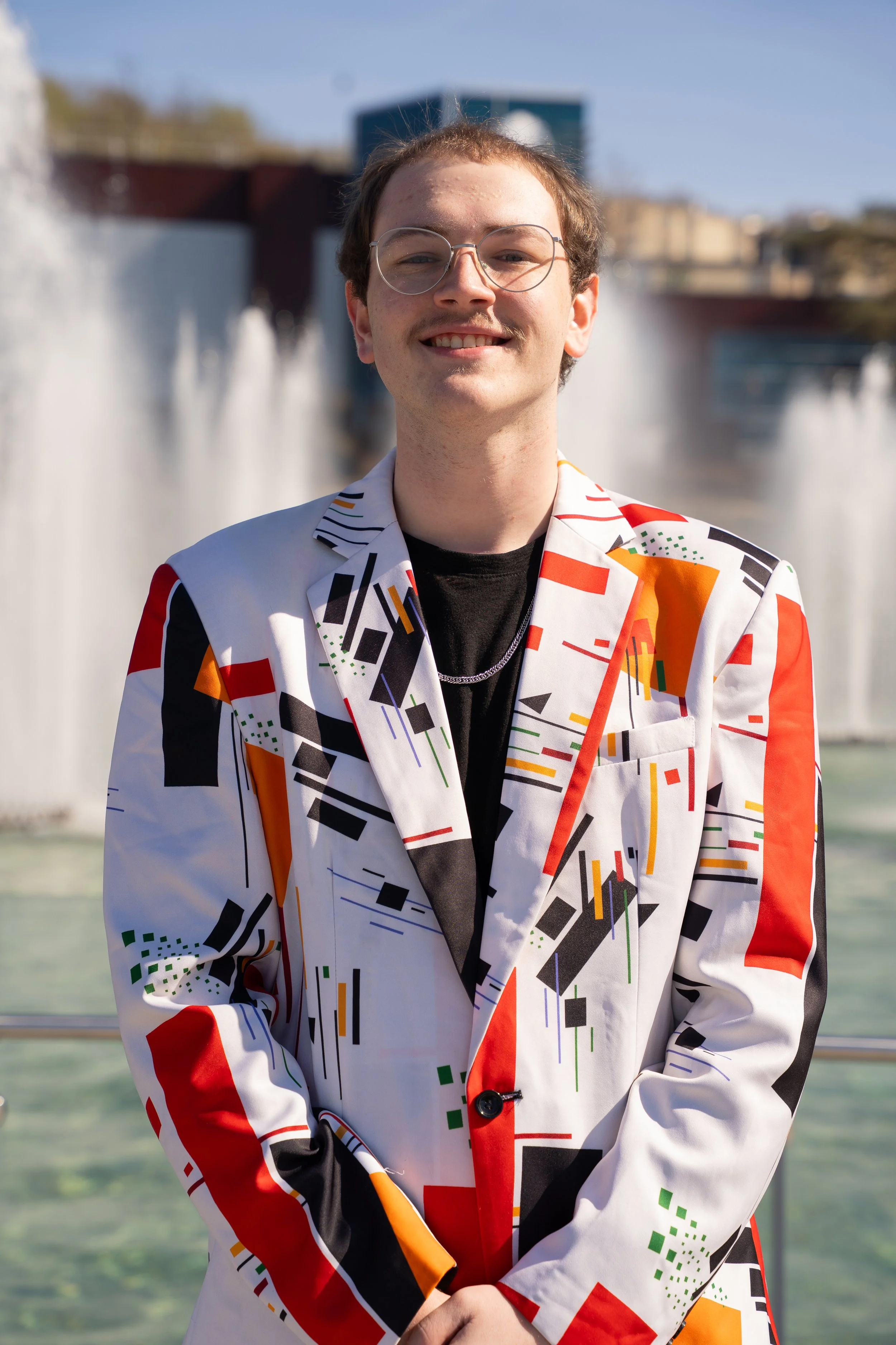 David Miller's Headshot, he is wearing a colorful blazer and standing in front of fountains.