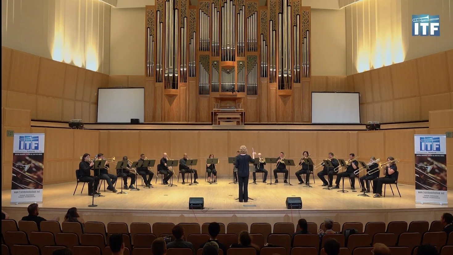 The 2023 Cramer Choir performing on stage in the Libby Gardner concert hall at the University of, directed by Dr. Natalie Mannix. There is a a large pipe organ in the background and two banners for the International Trombone Festival on either side.