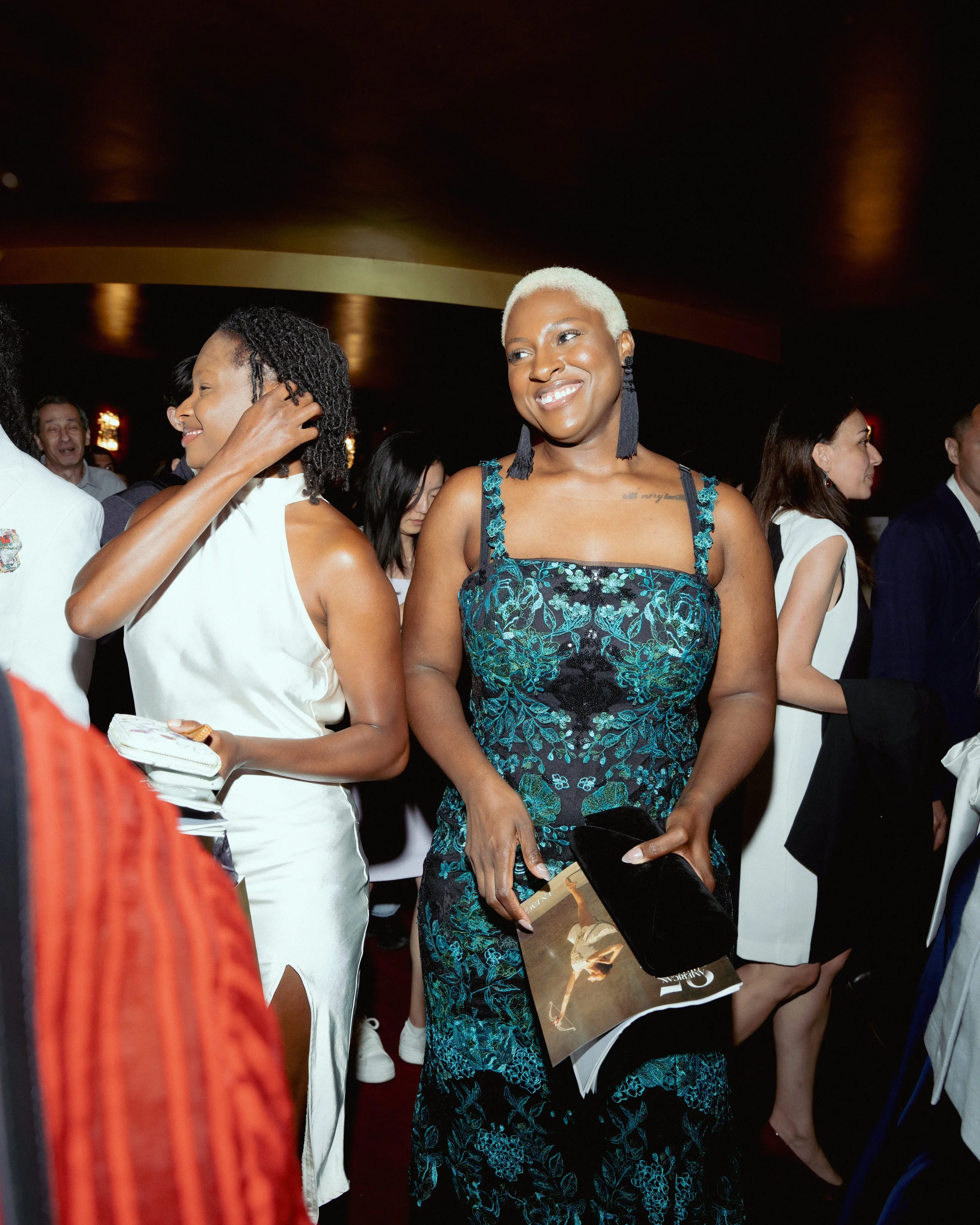 Group of people at a social event, with a woman with short platinum blonde hair and ornate teal and black dress in the foreground, smiling.