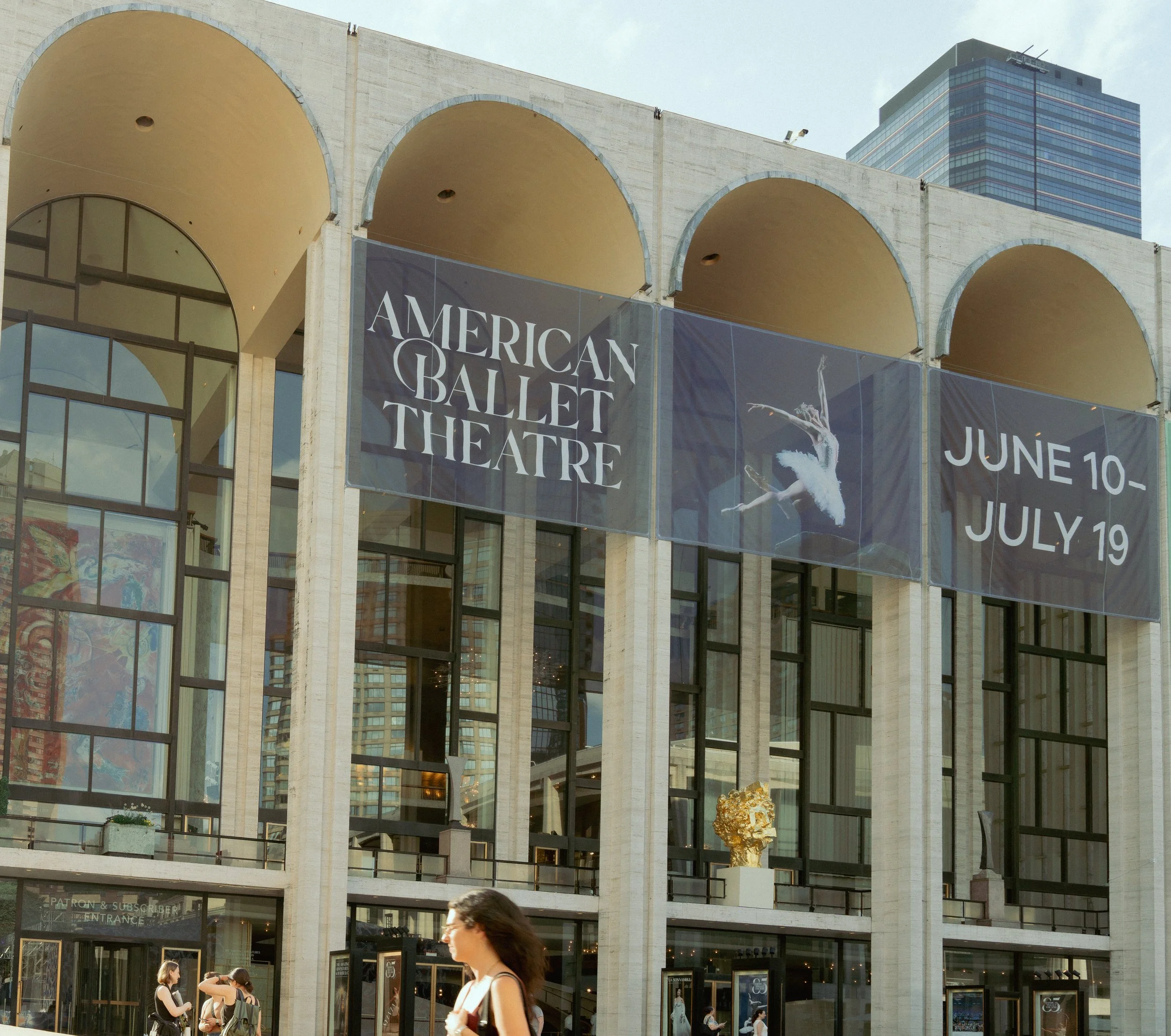 Exterior of the American Ballet Theatre building with large arches and glass windows, a banner advertising performances from June 10 to July 19, and people walking in front.