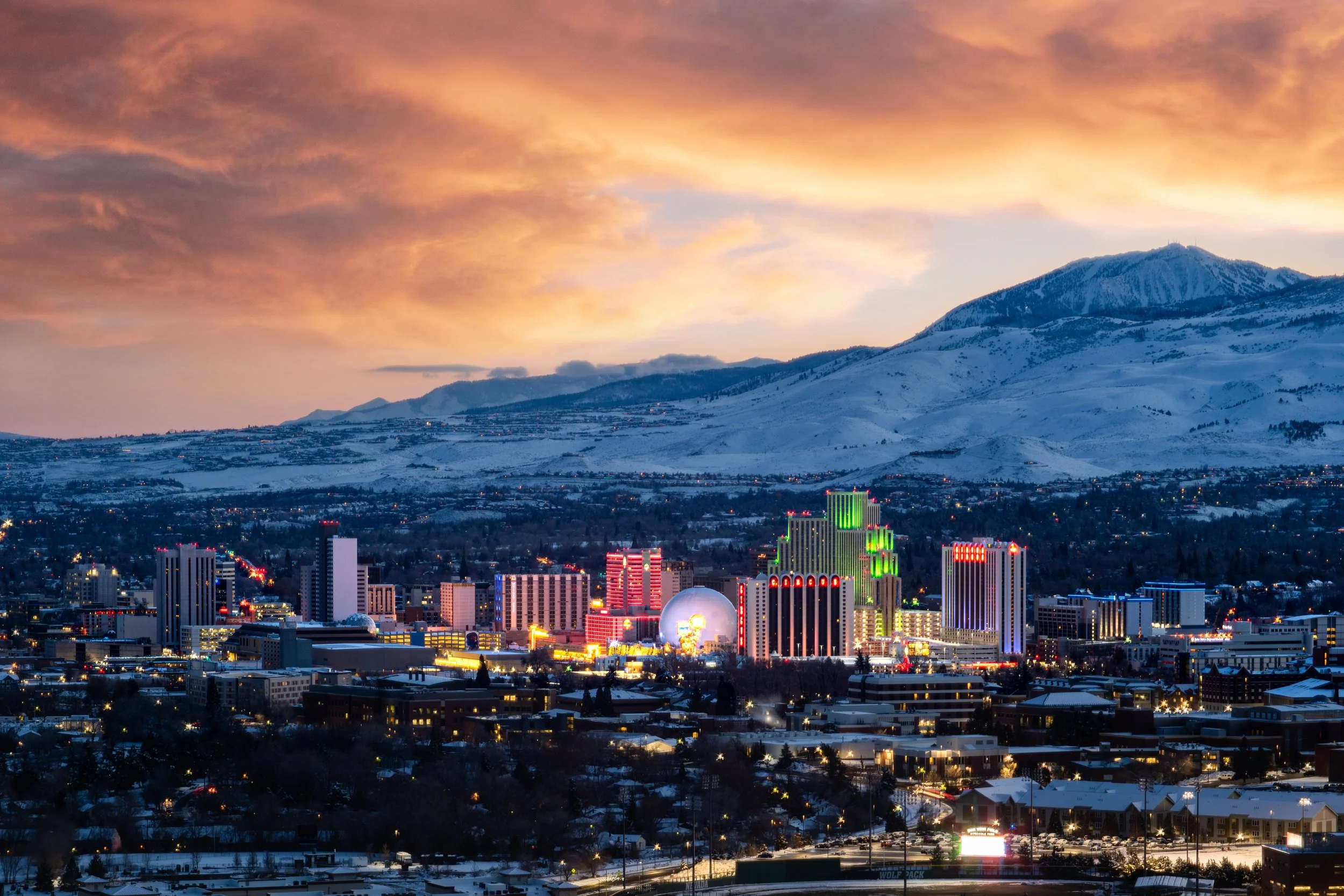 Nighttime cityscape with illuminated buildings and a snowy mountainous background at sunset.