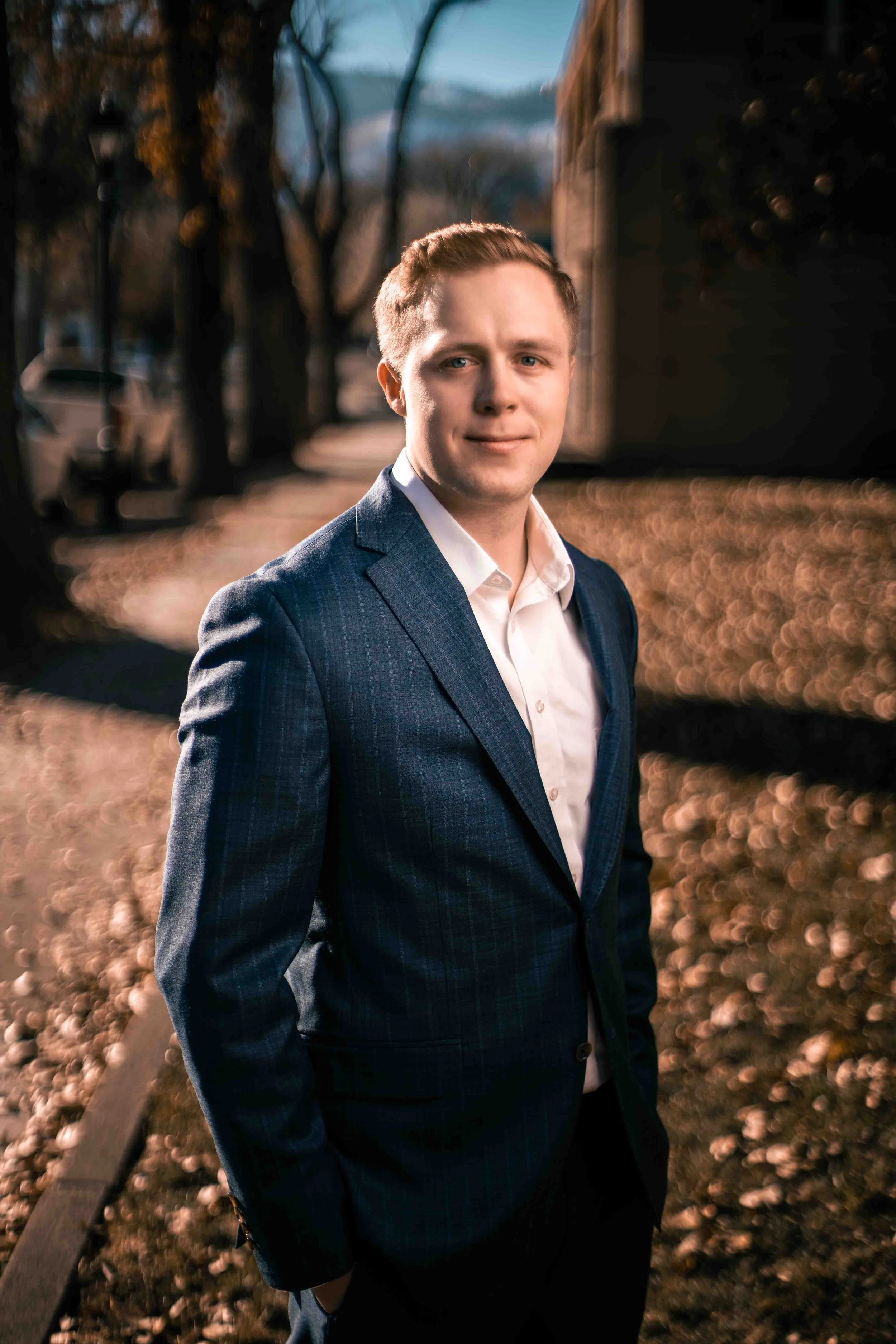 A young man with short light brown hair, wearing a dark blue checked suit and white shirt, standing on a sidewalk in a neighborhood during late afternoon with trees and a blurry house in the background.