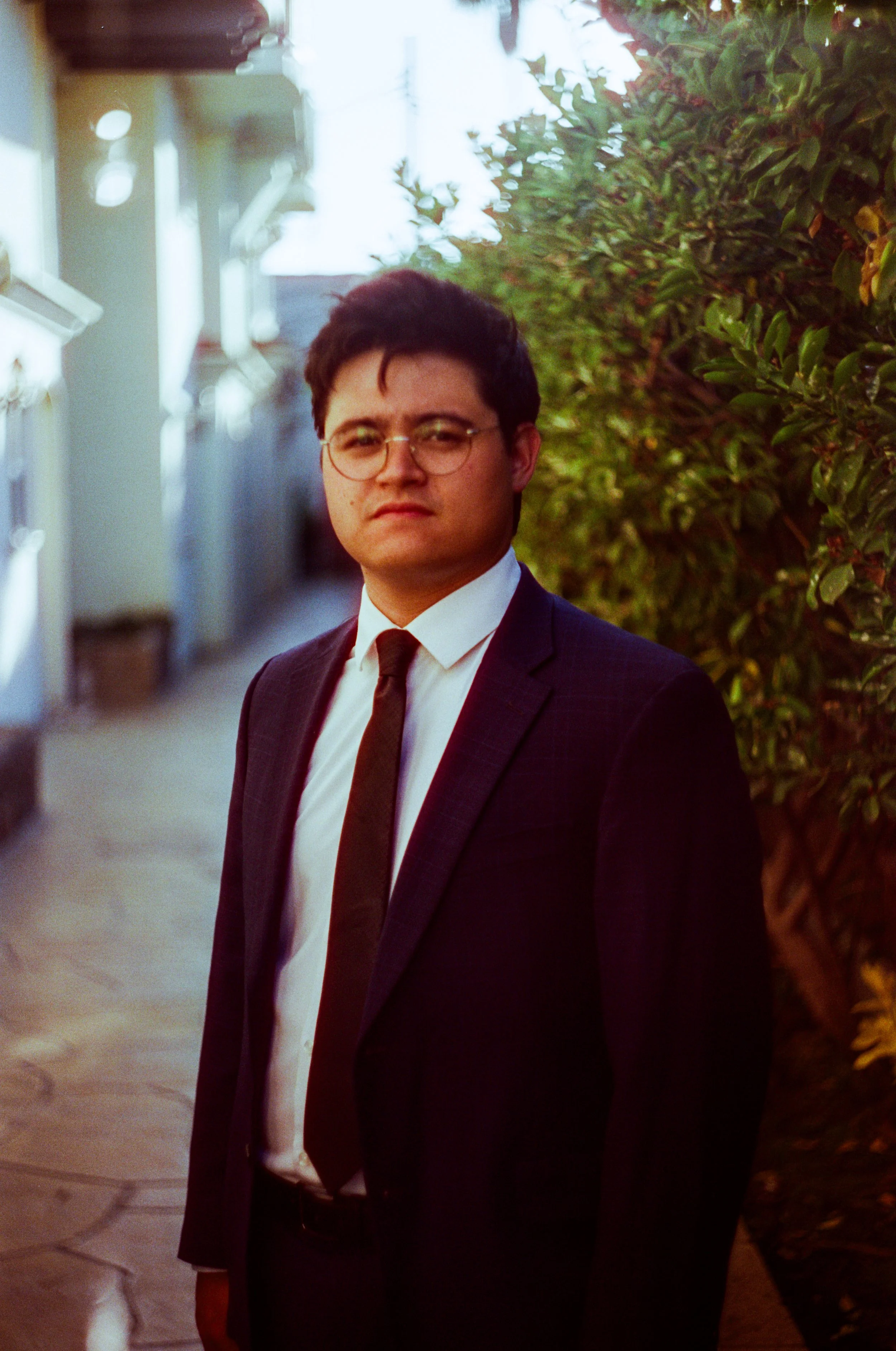 A young man in a suit and tie standing outdoors near a hedge, with houses in the background - Shot on 35mm film