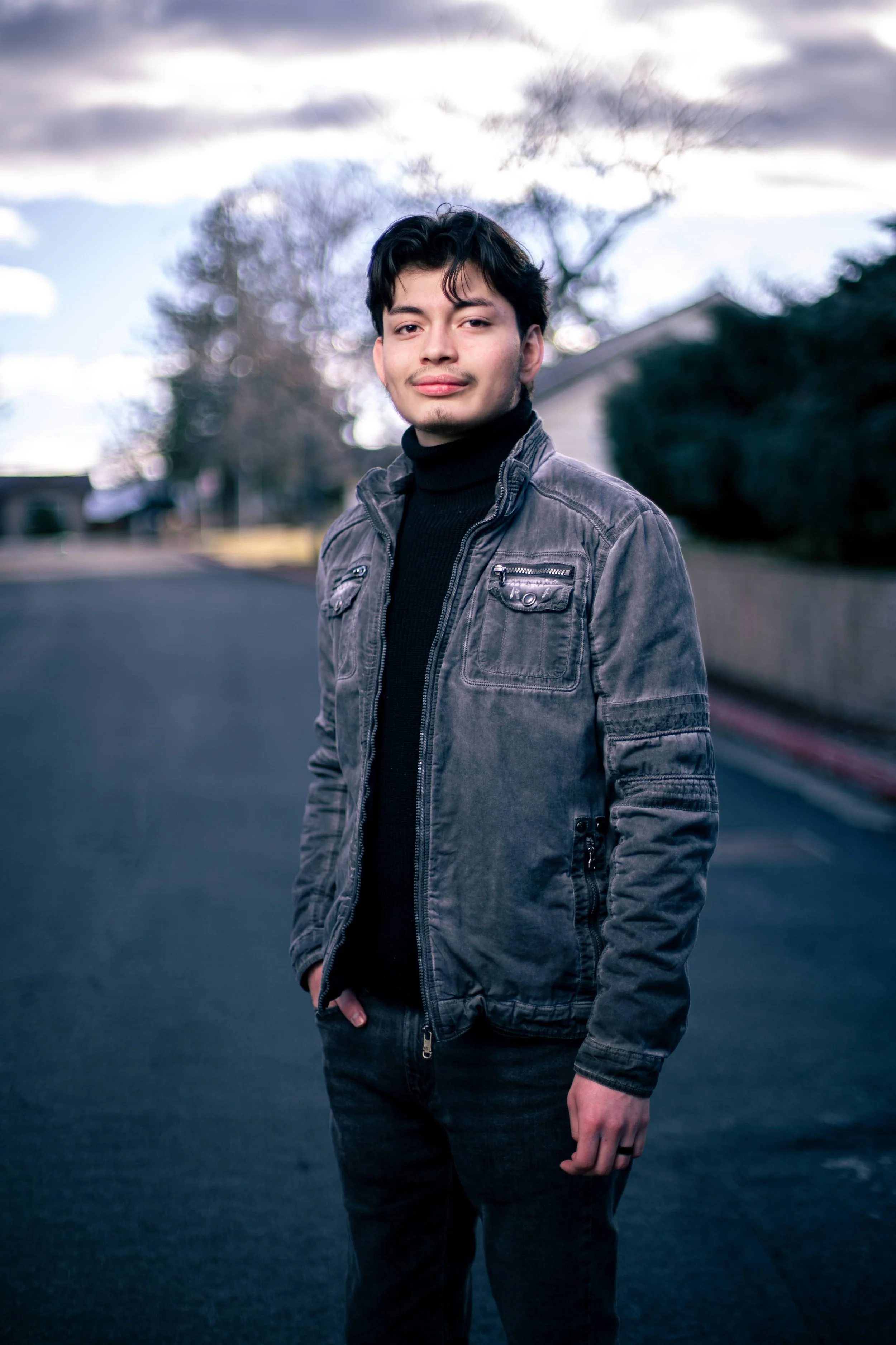 A young man standing outdoors on a paved street, wearing a black turtleneck sweater and gray jacket, with leafless trees and cloudy sky in the background.