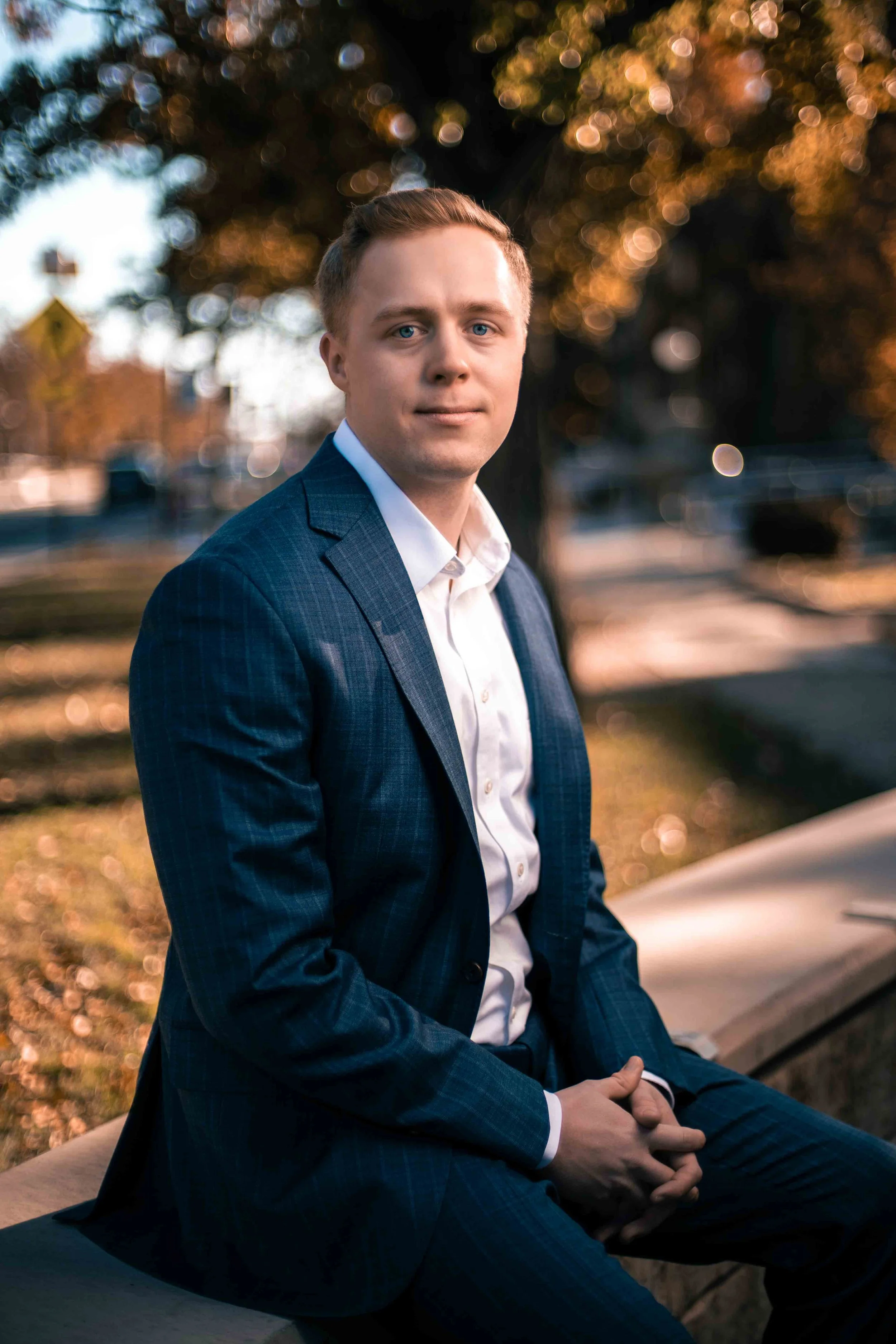 A young man in a blue suit and white shirt sitting outdoors near a tree with orange autumn leaves, during sunset, with a blurred background of a park and cars.