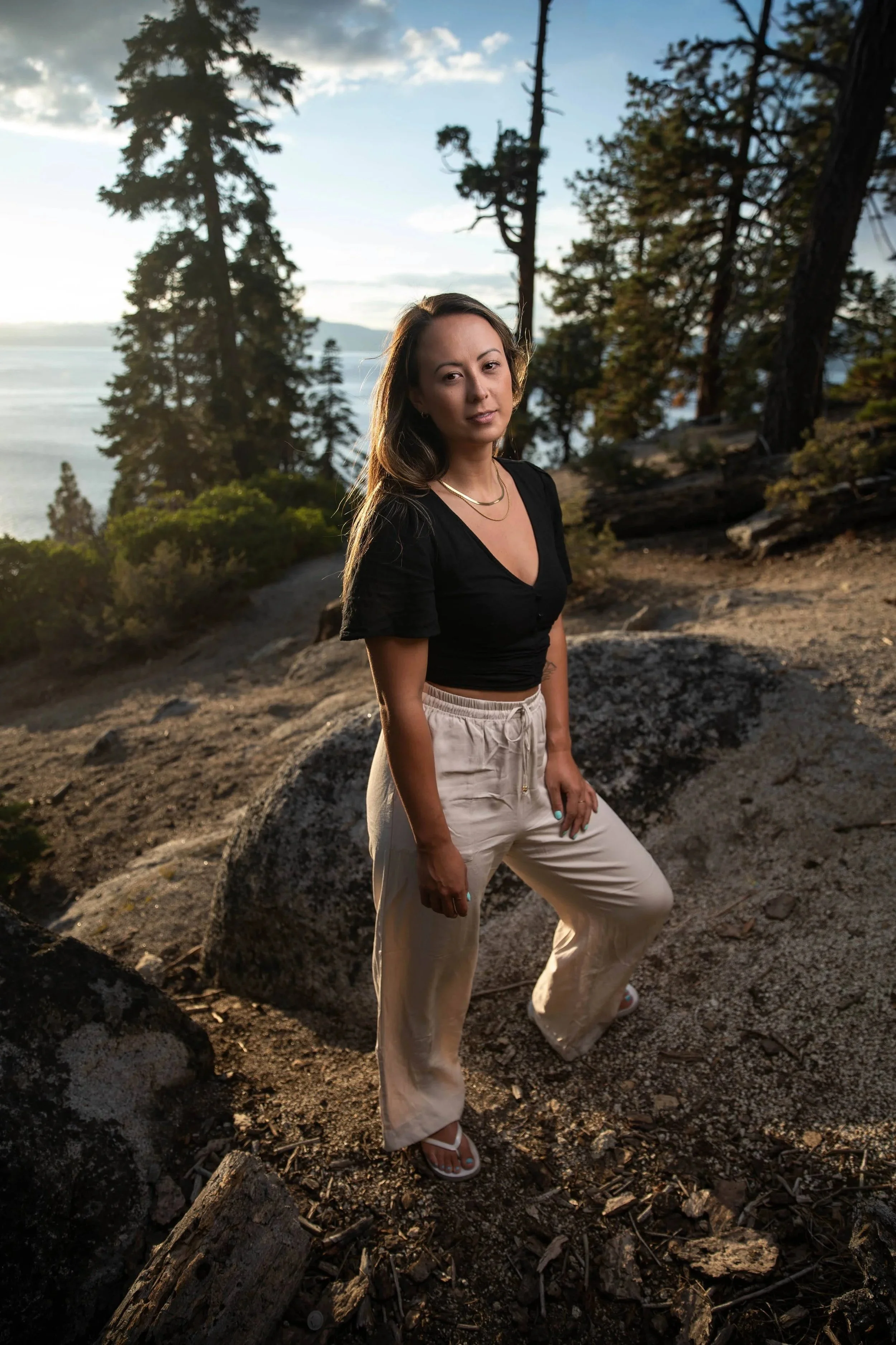 A woman standing outdoors on a rocky trail with trees and a body of water in the background during sunset.