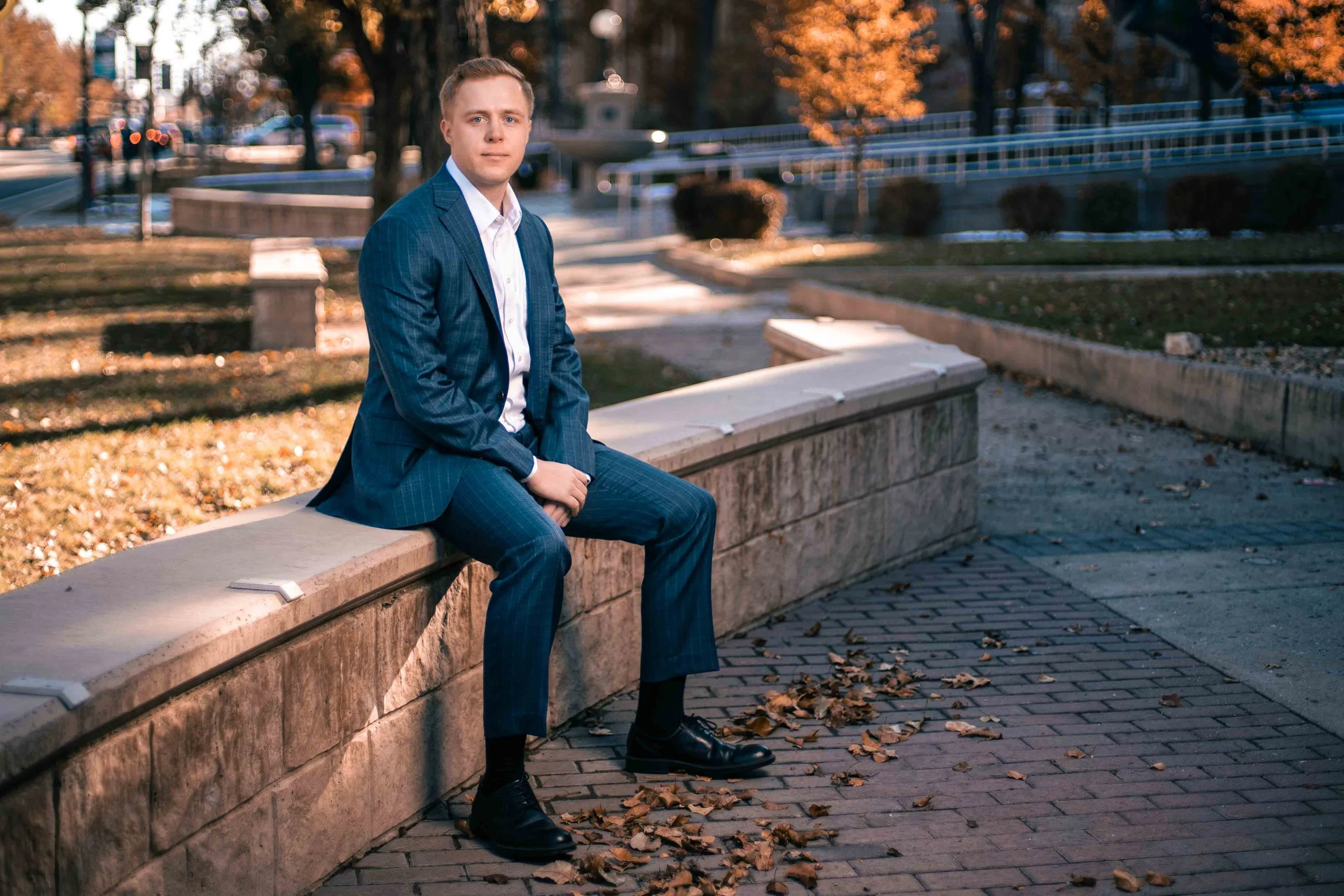 A young man in a blue suit sitting on a stone bench outdoors during autumn with fallen leaves around him, trees with orange leaves in the background, and a railing along the street.