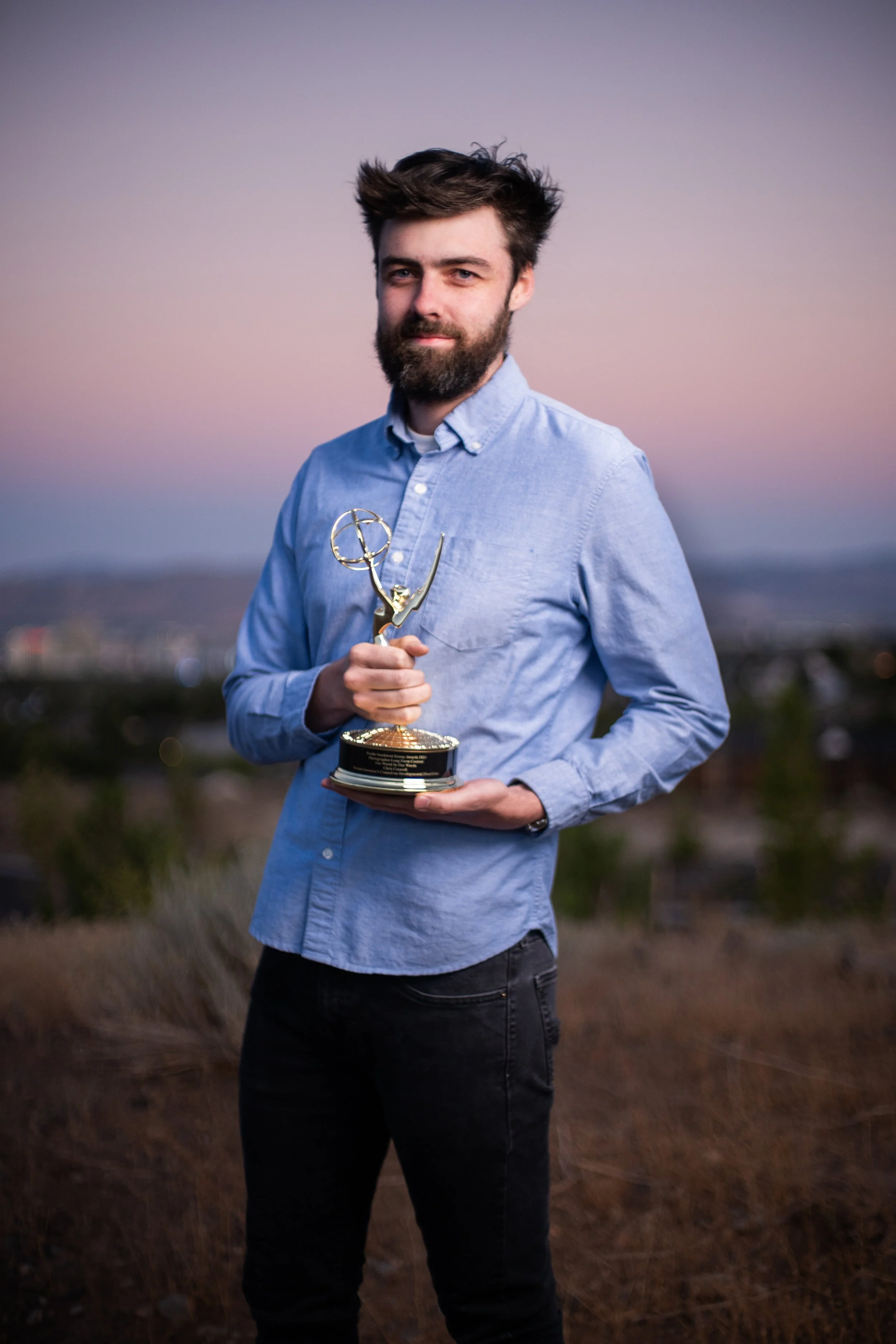 A man with a beard and short brown hair holding an Emmy award trophy outdoors during sunset, with a blurred landscape in the background.