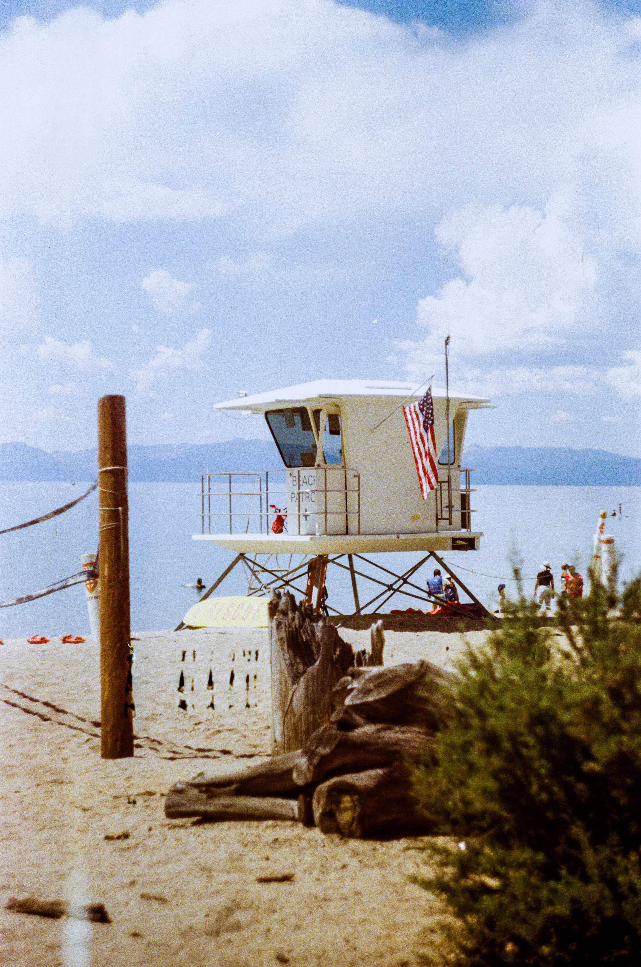 Lifeguard tower on a beach with people and water in the background, cloudy sky, and American flag hanging from the tower - Shot on 35mm film