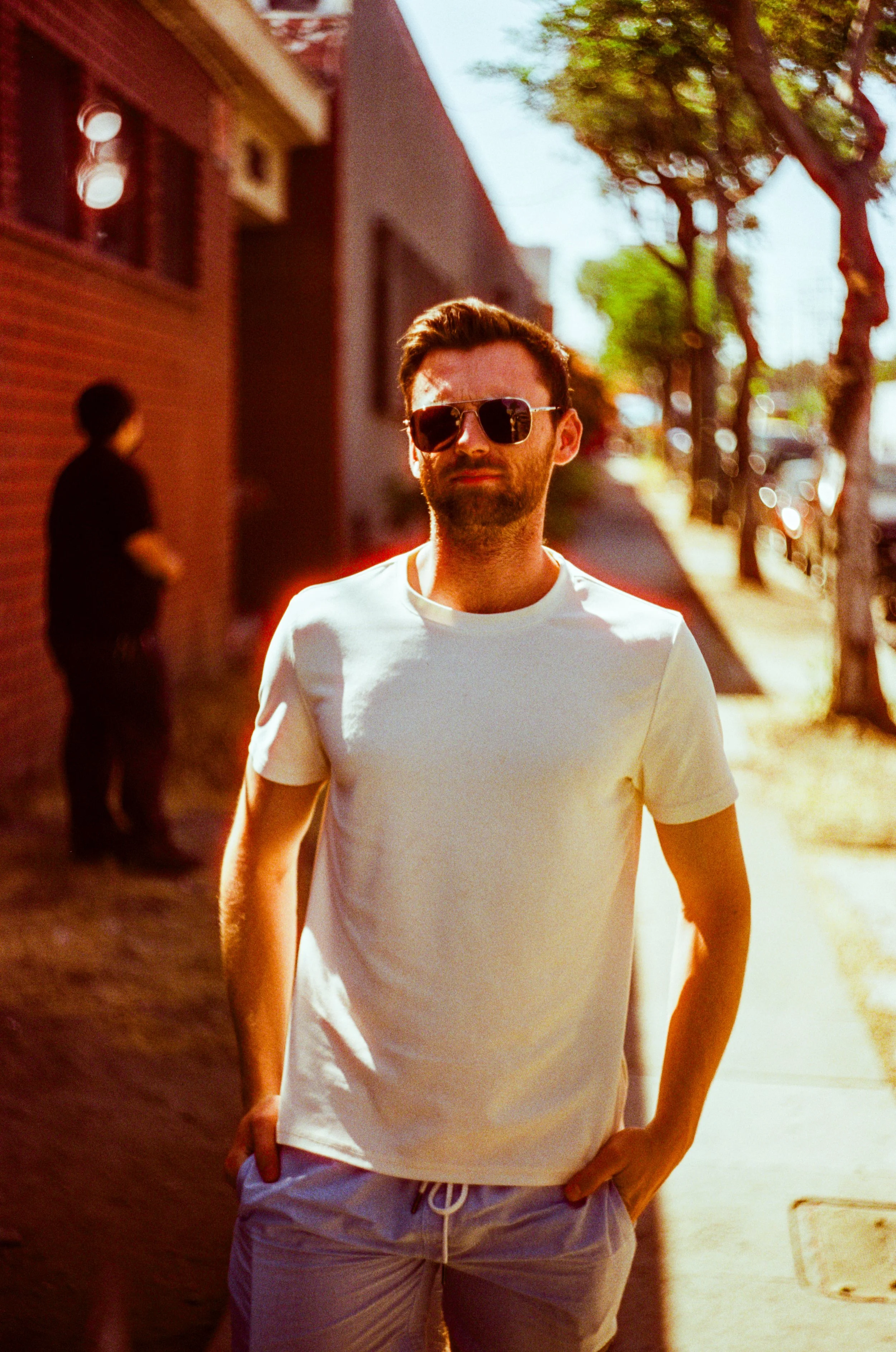 A man with a beard and sunglasses wearing a white t-shirt and shorts standing on a sidewalk in a city, with trees and buildings in the background - Shot on 35mm film