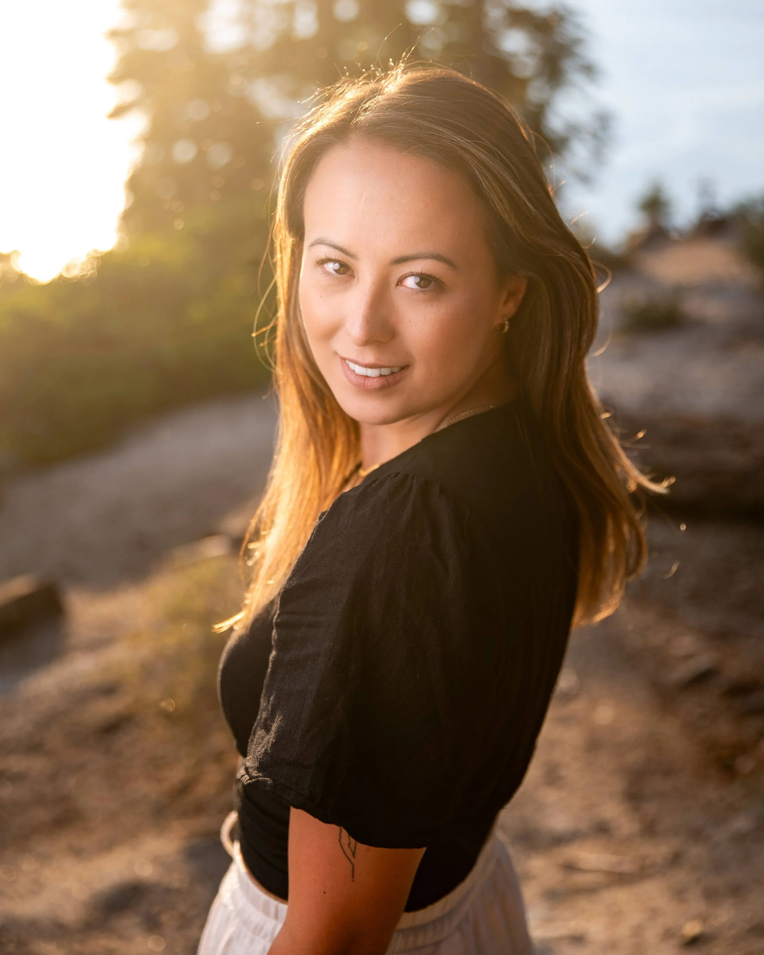 Young woman with brown hair smiling outdoors during sunset, wearing a black top and light-colored skirt, with blurred trees and sky in the background.