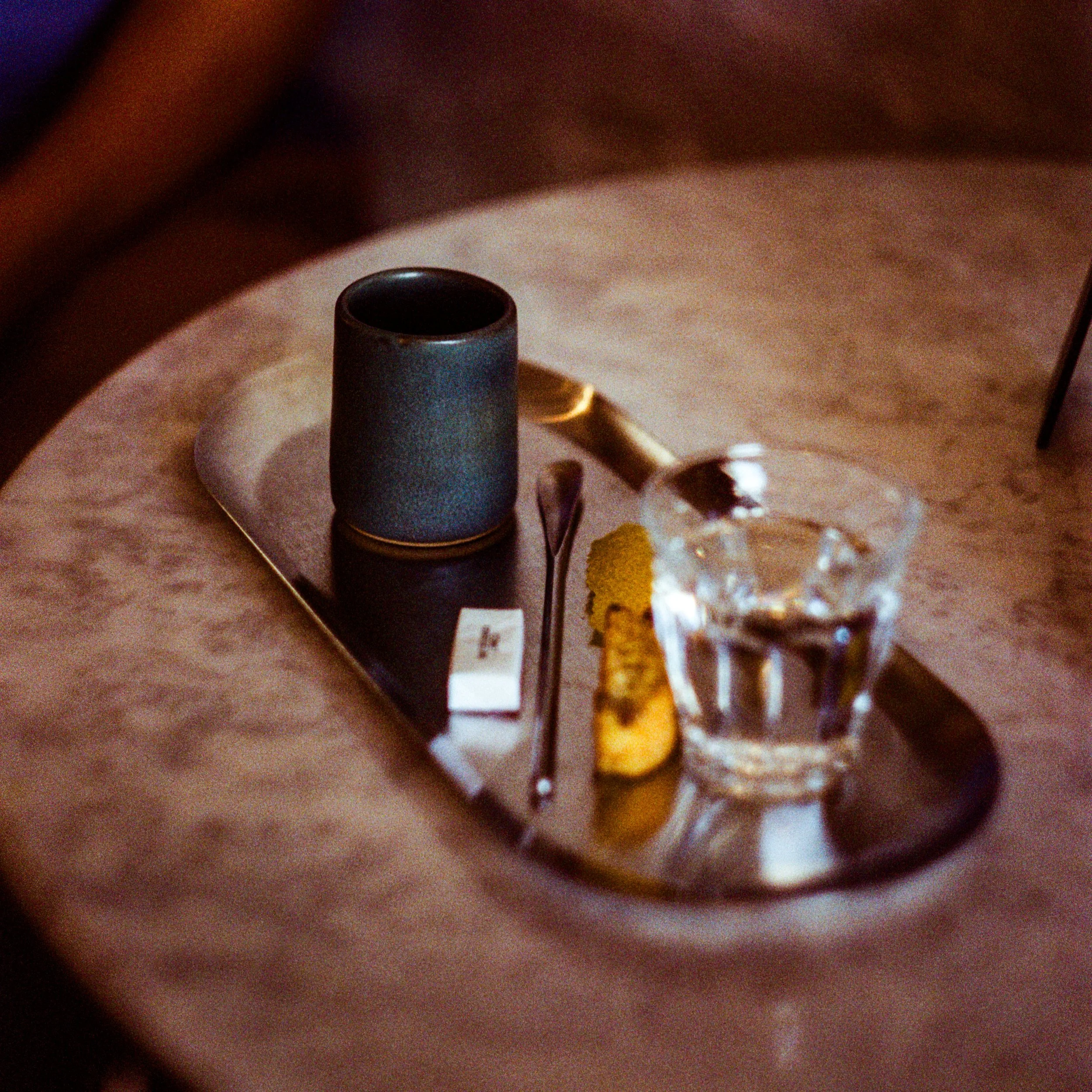 A metal tray on a marble table with a dark ceramic cup, a spoon, a small bowl of sugar, and a glass of water - Shot on 35mm film