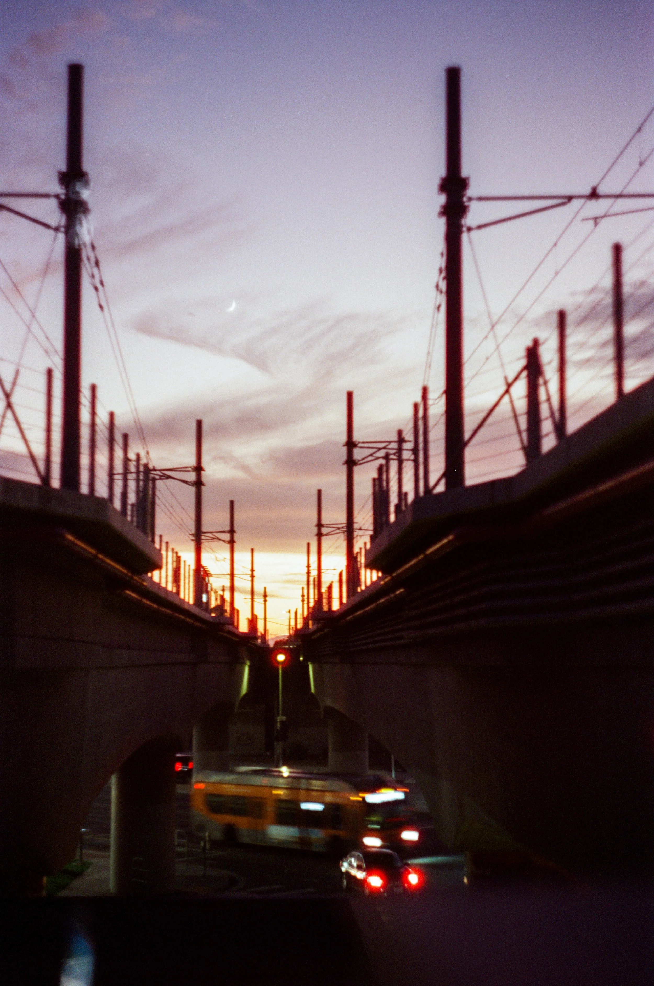 Photo of a street scene at sunset with a bridge, cars, and a bus, and a visible crescent moon in the sky.