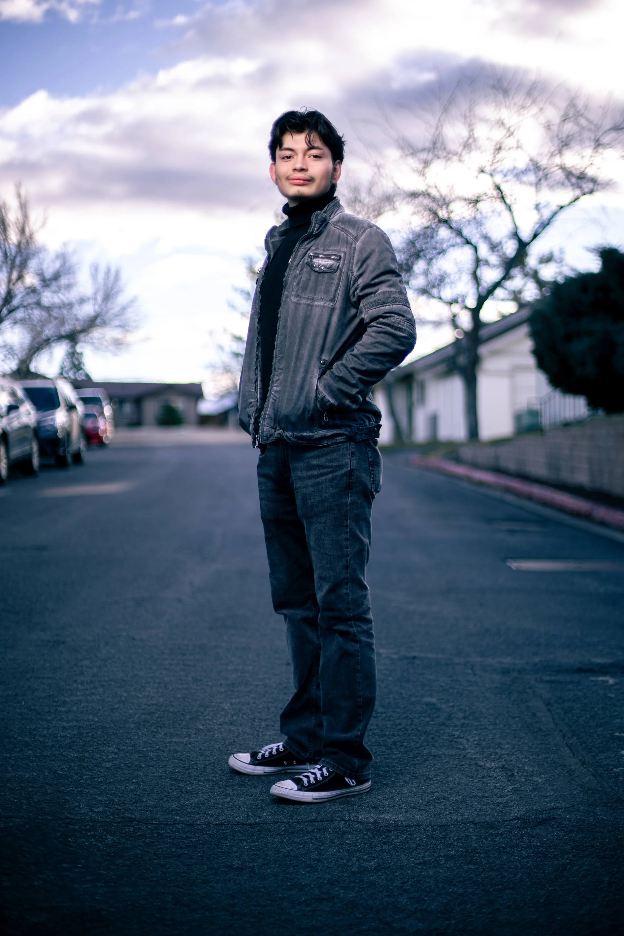 A young man standing in an outdoor parking lot with parked cars and trees in the background, wearing a black jacket, jeans, and Converse sneakers, during a cloudy day.