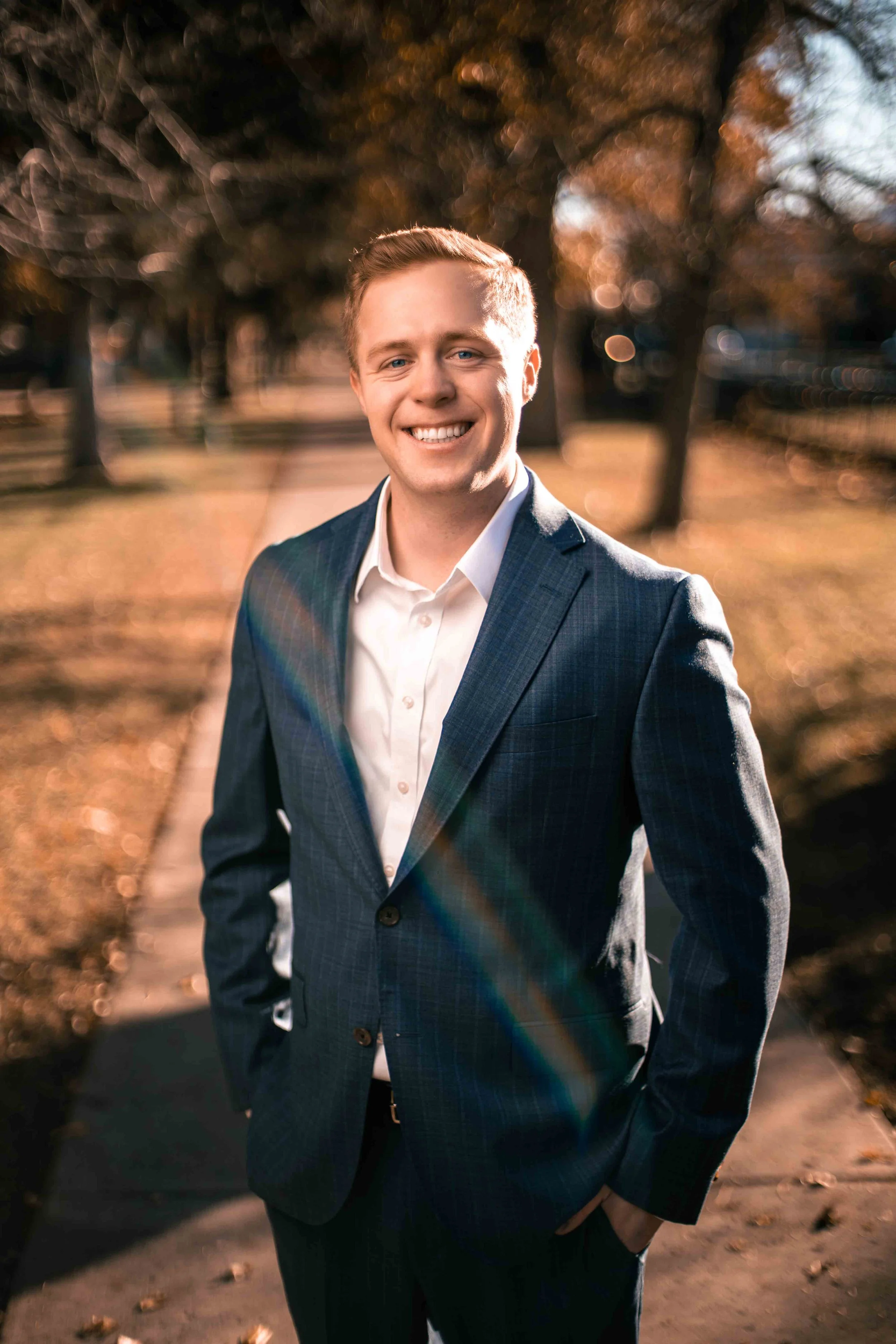 A young man with blonde hair smiling outdoors in a suit and white shirt, with a park and trees in the background, during late afternoon sunlight.