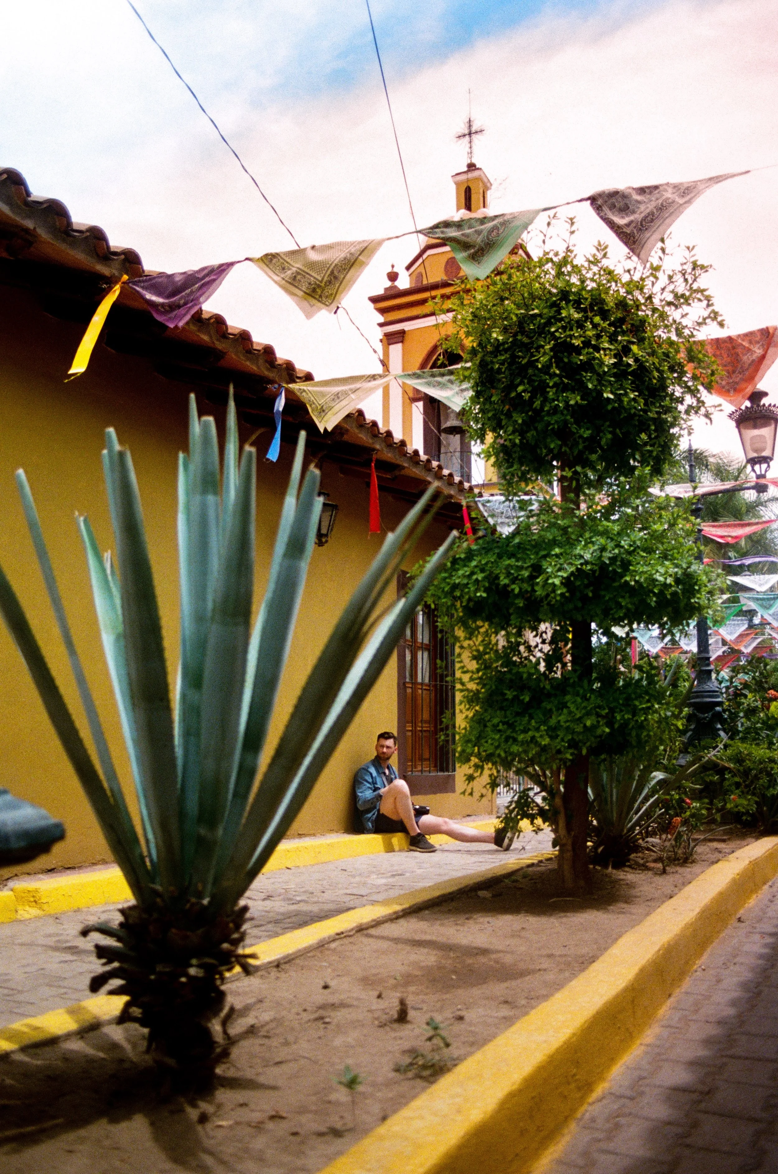 A street scene with colorful flags hanging overhead, a yellow building with a bell tower in the background, and a man sitting on the sidewalk near a tree, with plants and lanterns lining the street.