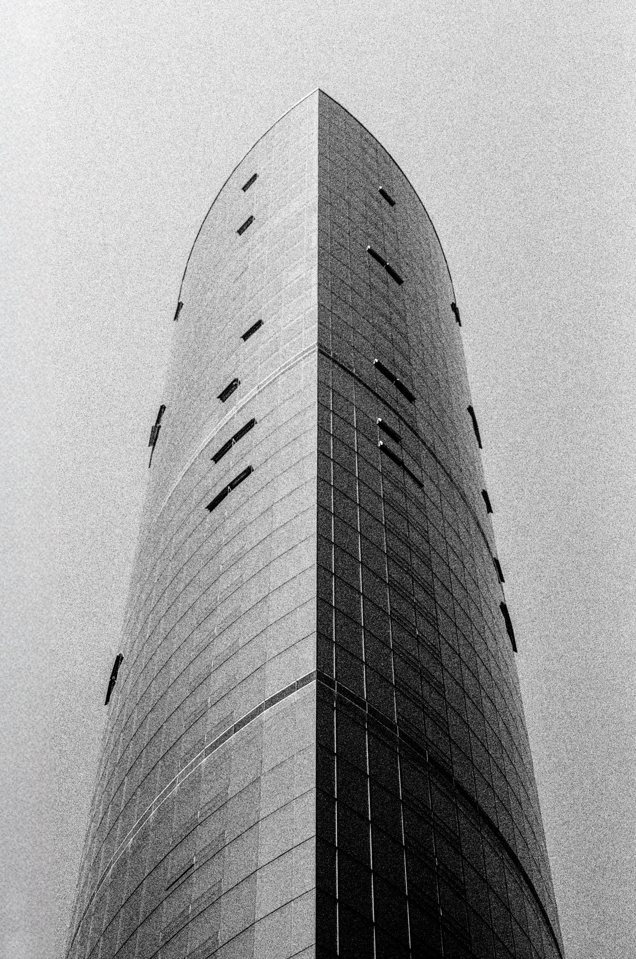 Black and white photo of a tall, curved modern skyscraper viewed from below against a gray sky - Shot on 35mm film