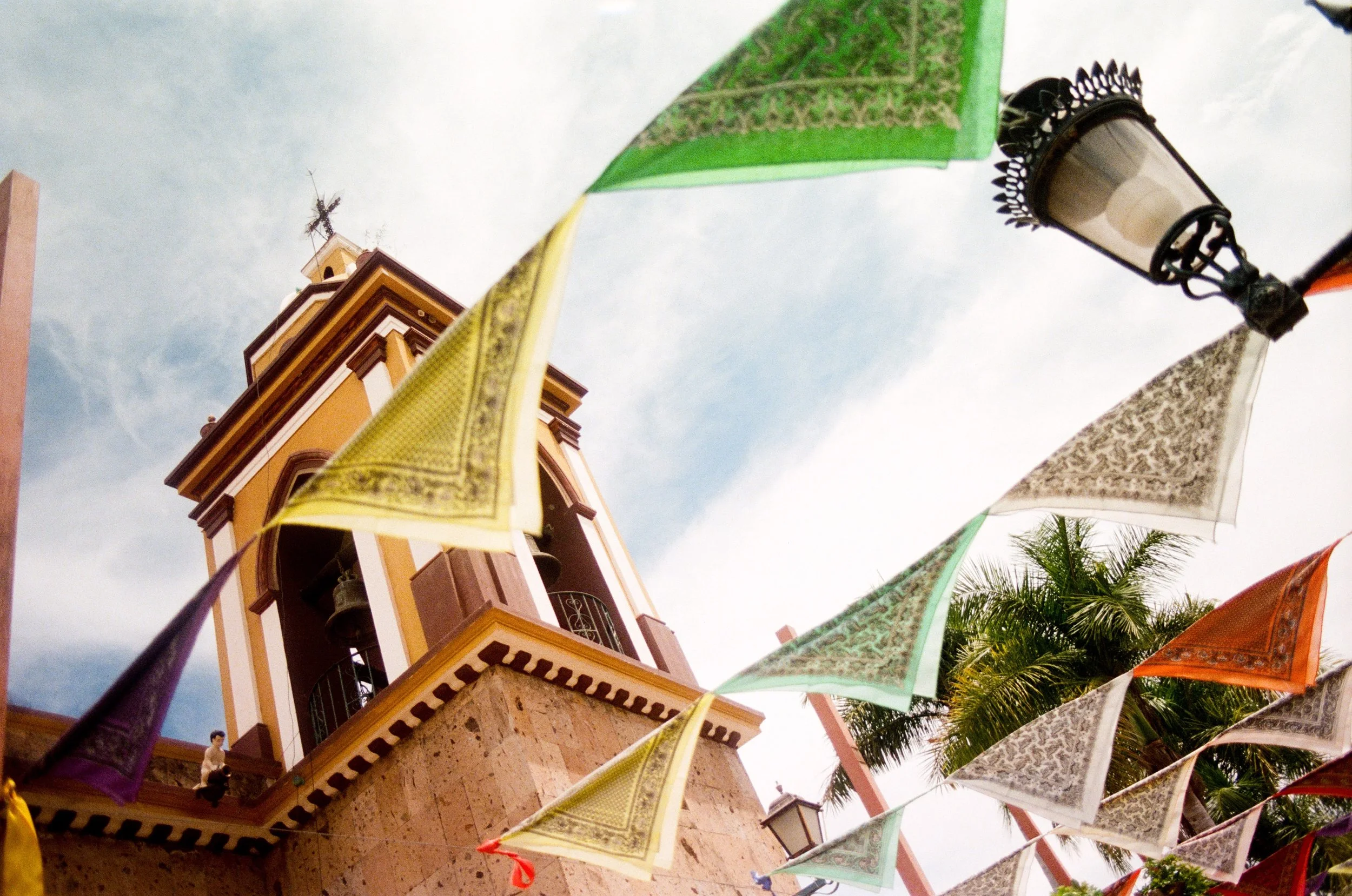 Colorful decorative flags and lanterns hang above a yellow church steeple with a cross on top. The sky is partly cloudy, and there are palm trees in the background.