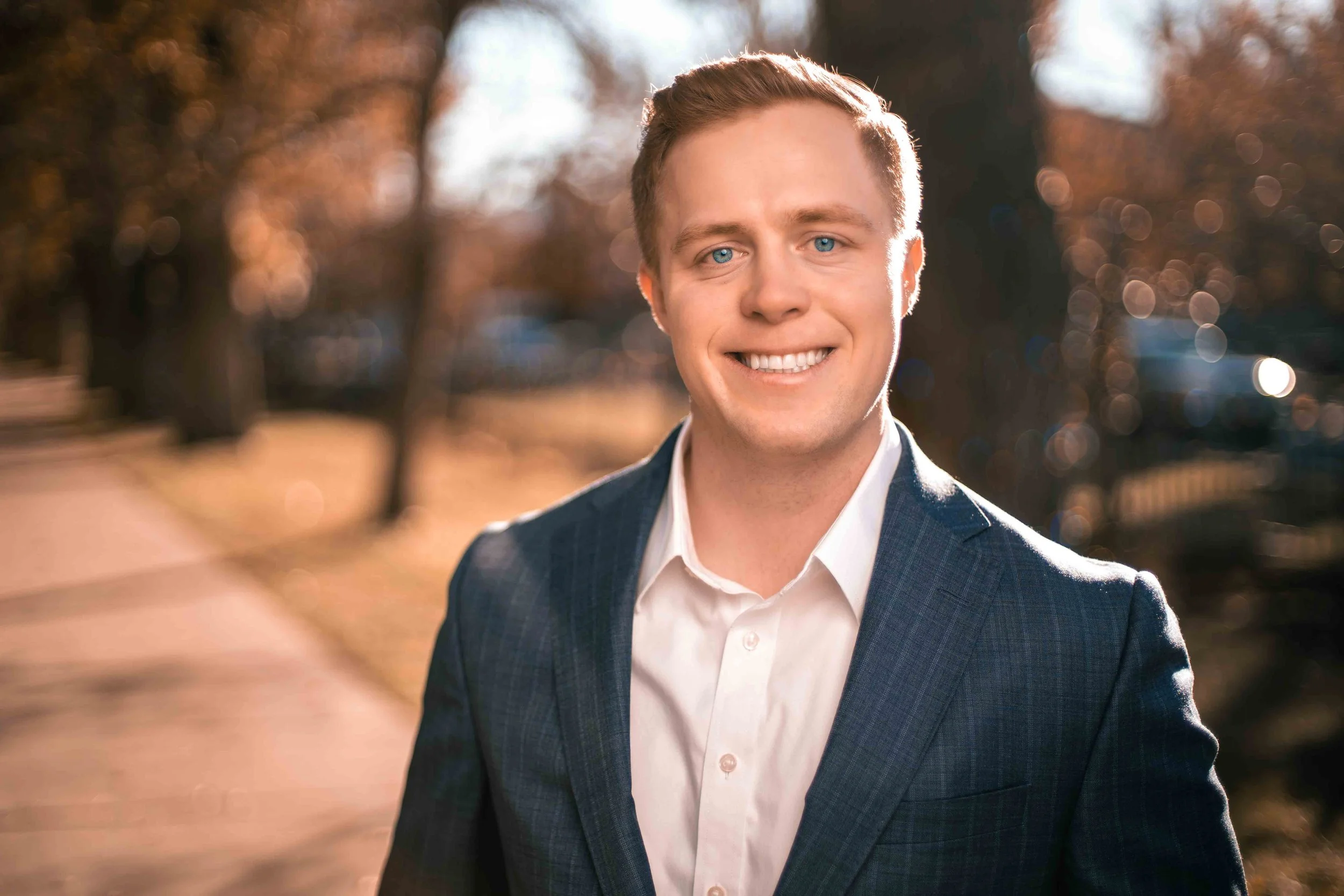 A young man wearing a white shirt and dark blue blazer smiling outdoors on a sunny day with trees and a sidewalk in the background.