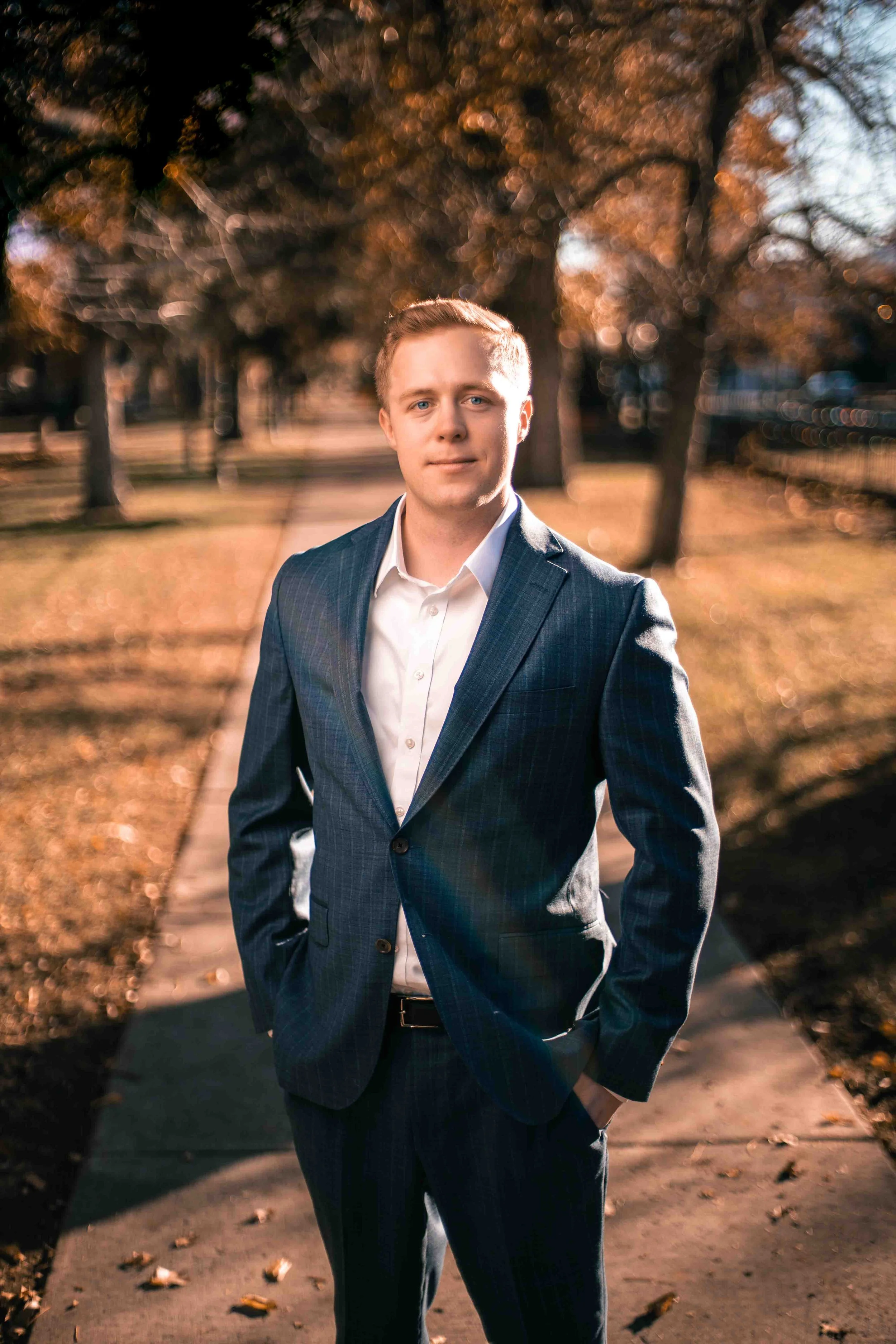 A young man dressed in a suit and white shirt standing on a sidewalk in a park during fall, with trees in the background and sunlight casting warm tones.