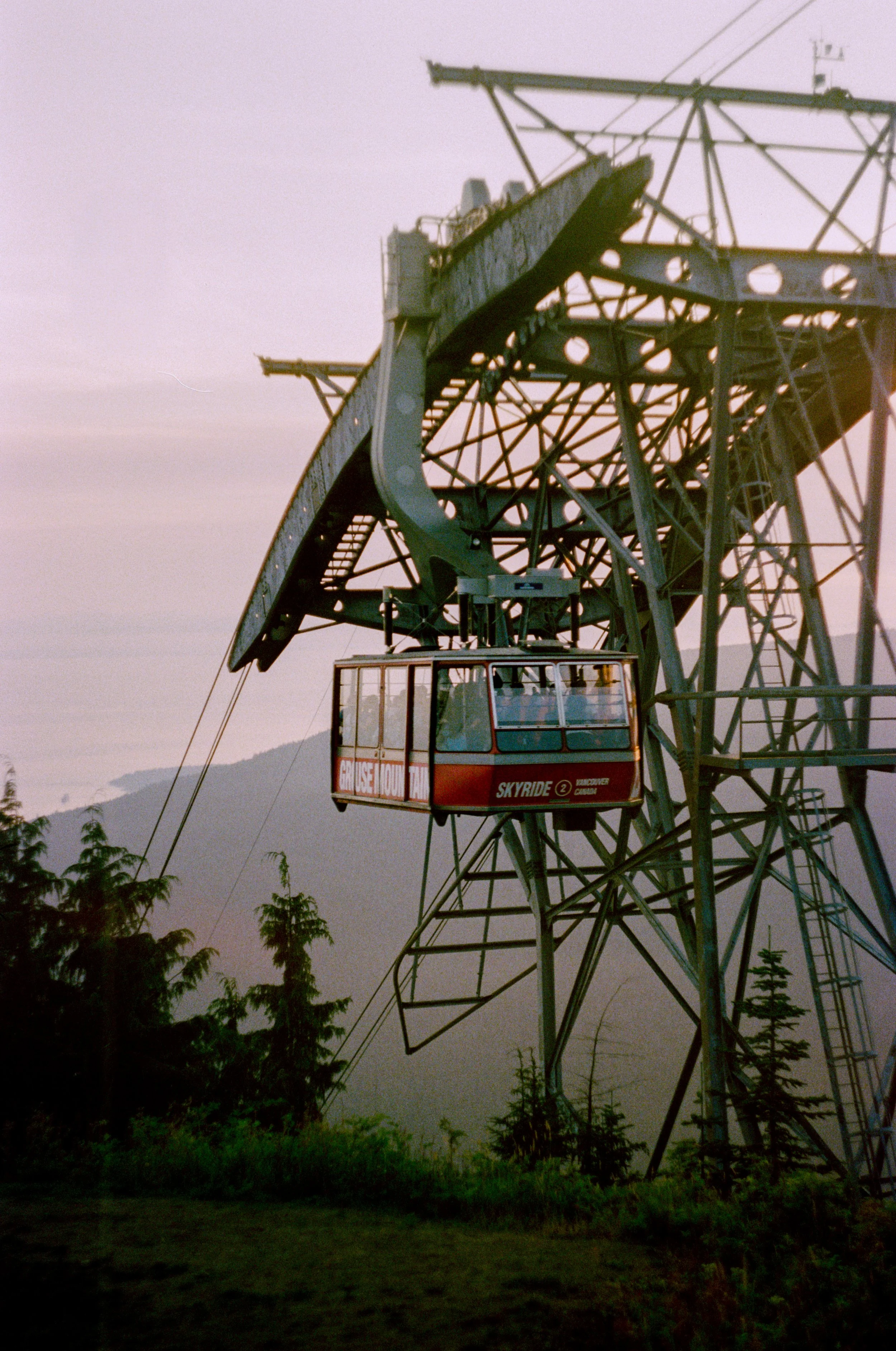Grouse Mountain Tram - featuring a red cable car hanging from a large green metal structure, with a backdrop of distant mountains and trees - Shot on 35mm film