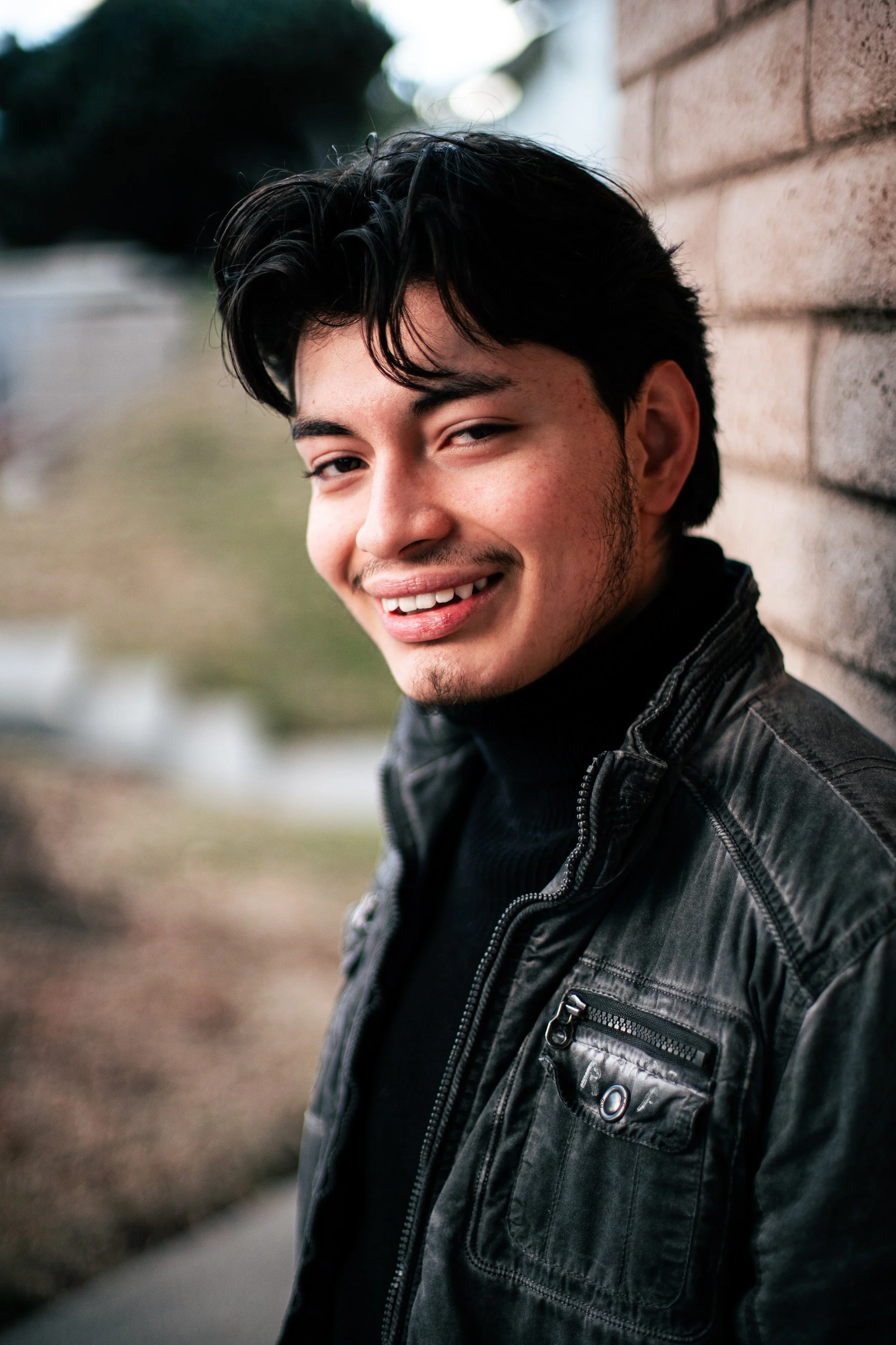 A young man with dark hair, a slight smile, and facial hair, wearing a black leather jacket and a black turtleneck, leaning against a brick wall outdoors with blurred background.