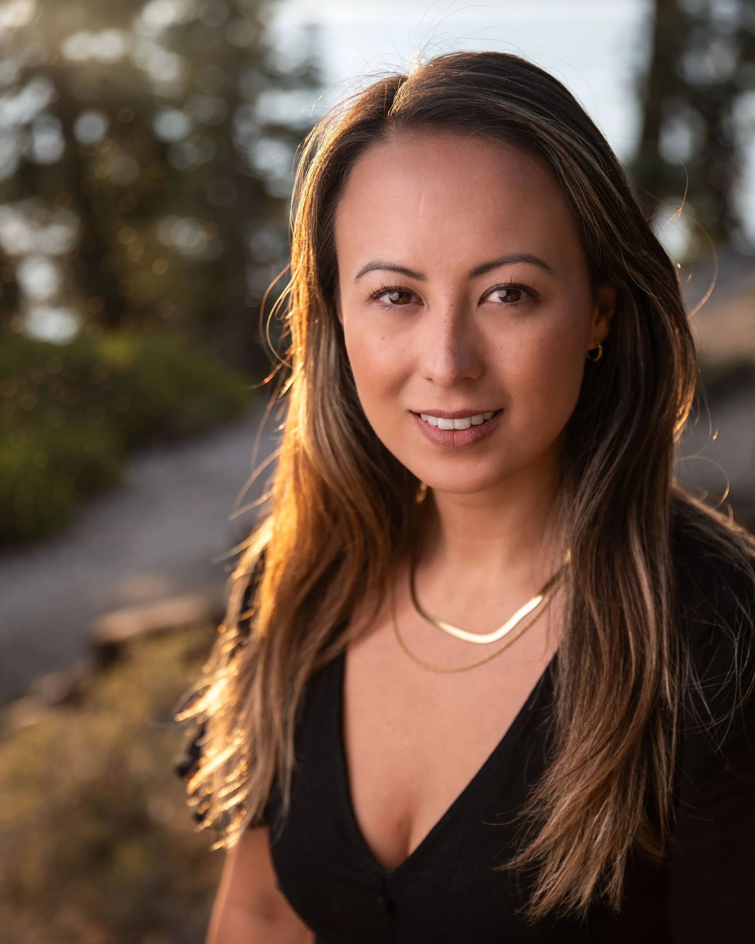 A woman with long brown hair outdoors during sunset, wearing a black top, gold necklaces, and small earrings, smiling softly at the camera.