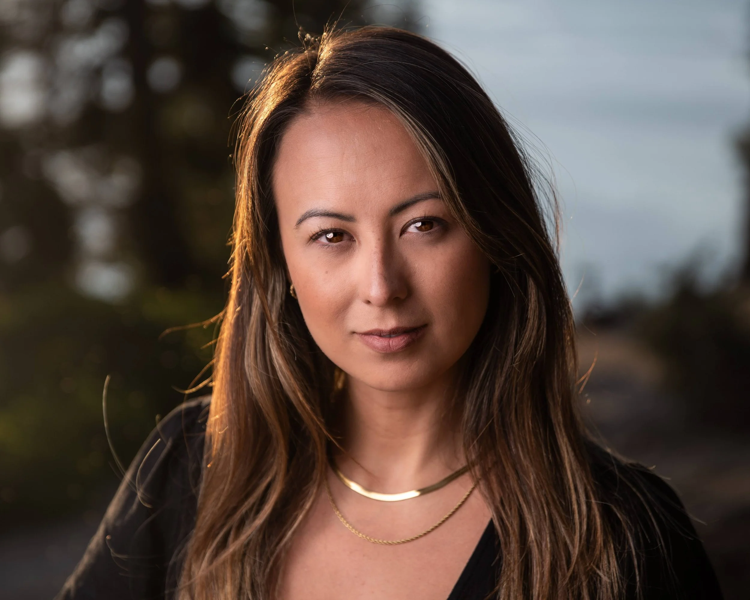 A woman with long brown hair, wearing a black top and gold jewelry, posing outdoors during sunset with a blurred natural background.