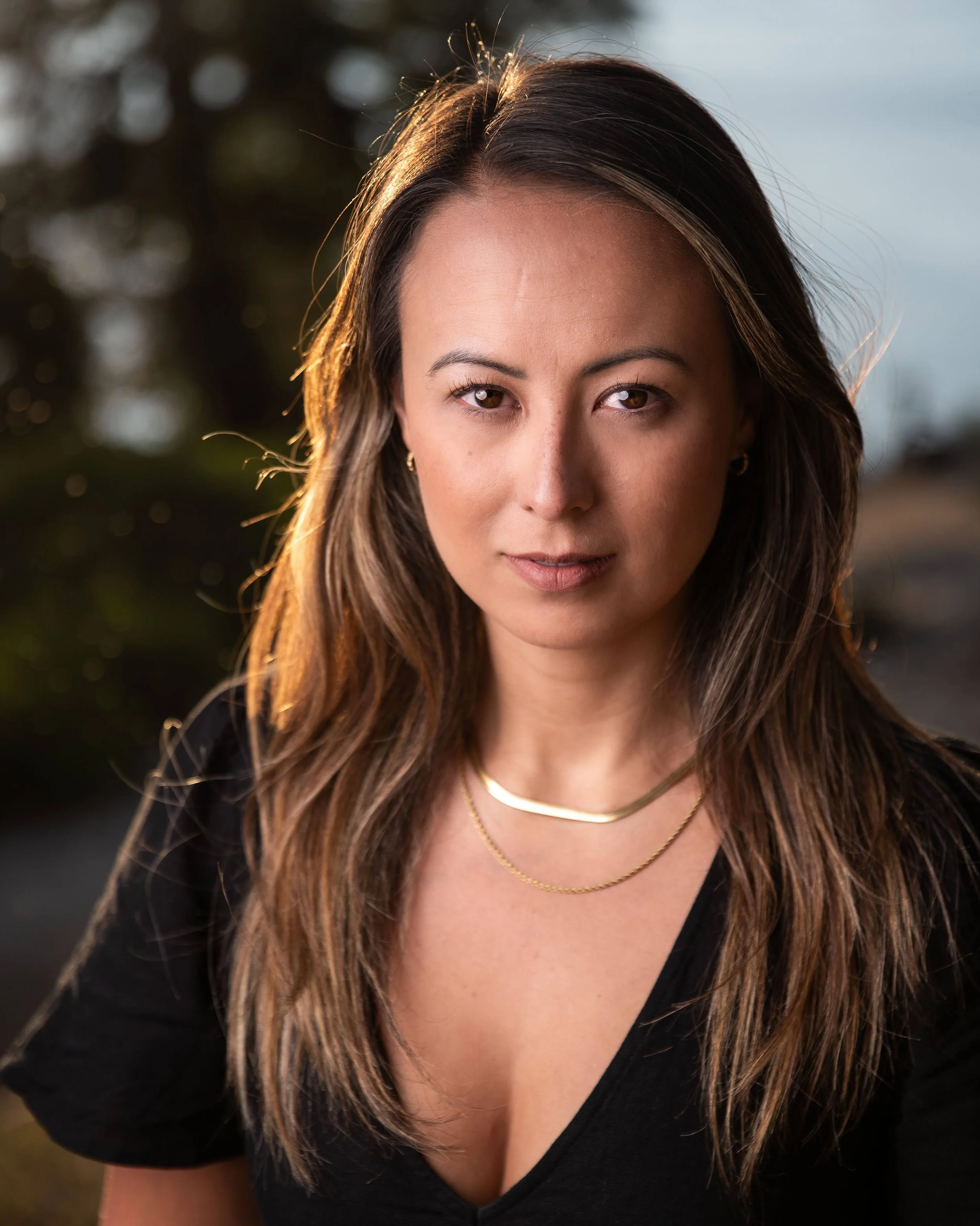 A woman with long brown hair, wearing a black top and layered necklaces, posing outdoors during golden hour with a blurred background of trees and sky.