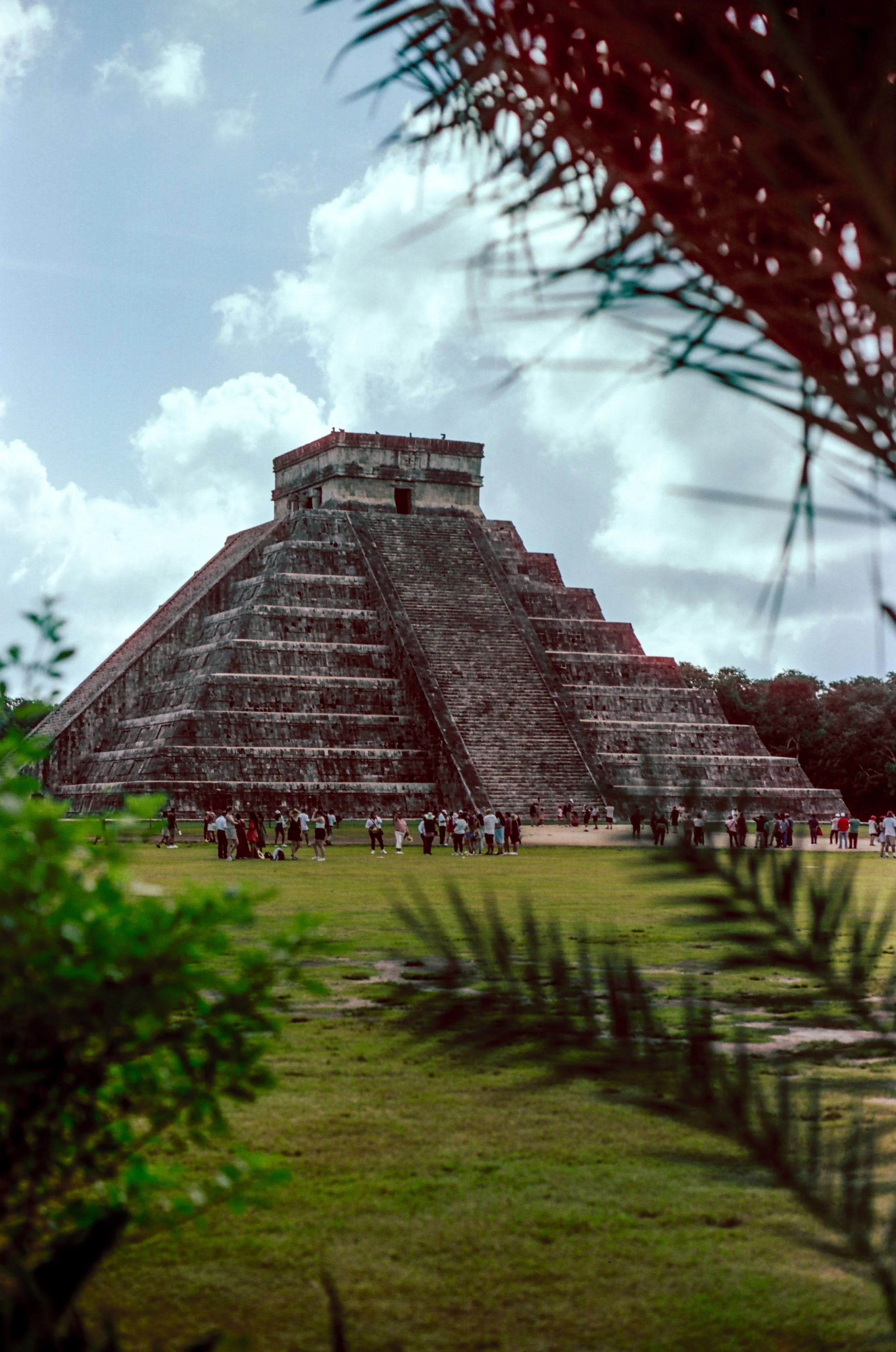 Ancient Mayan pyramid at Chichen Itza, viewed through tropical foliage with a blue sky and clouds in the background, and a crowd of tourists at its base - Shot on 35mm film