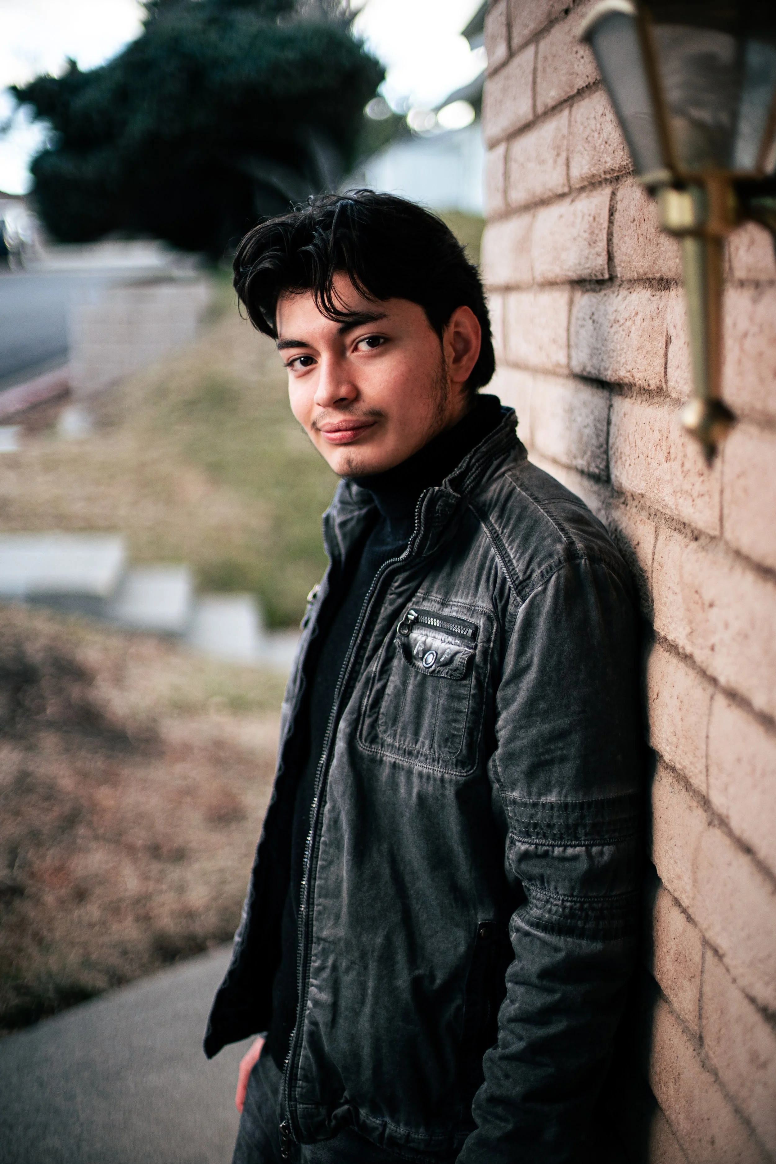 A young man with black hair and light skin leaning against a brick wall outdoors, wearing a black leather jacket and looking at the camera.