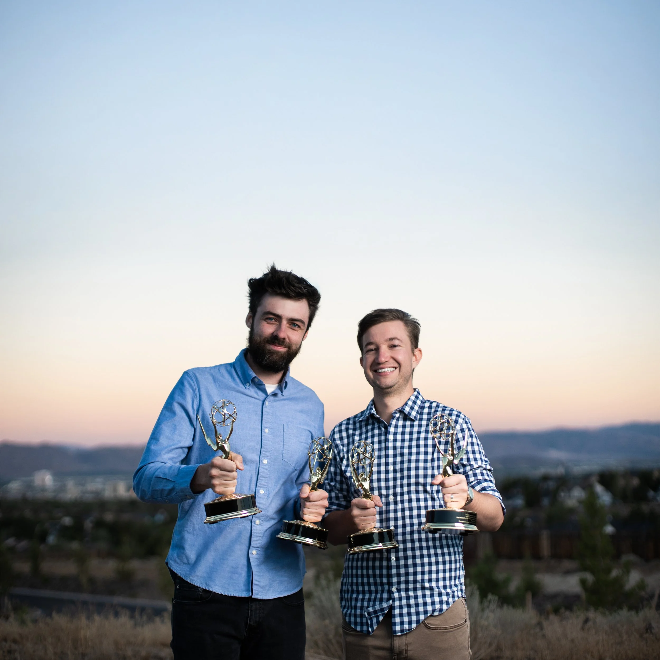 Two young men standing outdoors at sunset, holding Emmy awards and smiling.