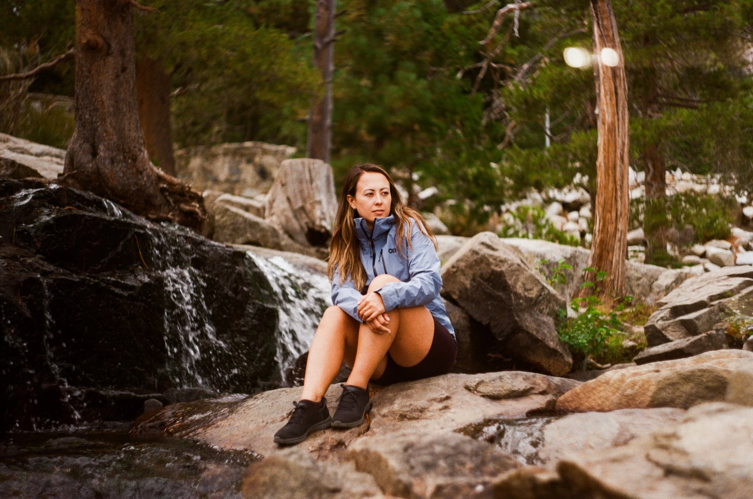 A woman sitting on rocks in a forest near a small waterfall, wearing a blue jacket and black shoes, with trees and rocks in the background  - Shot on 35mm film
