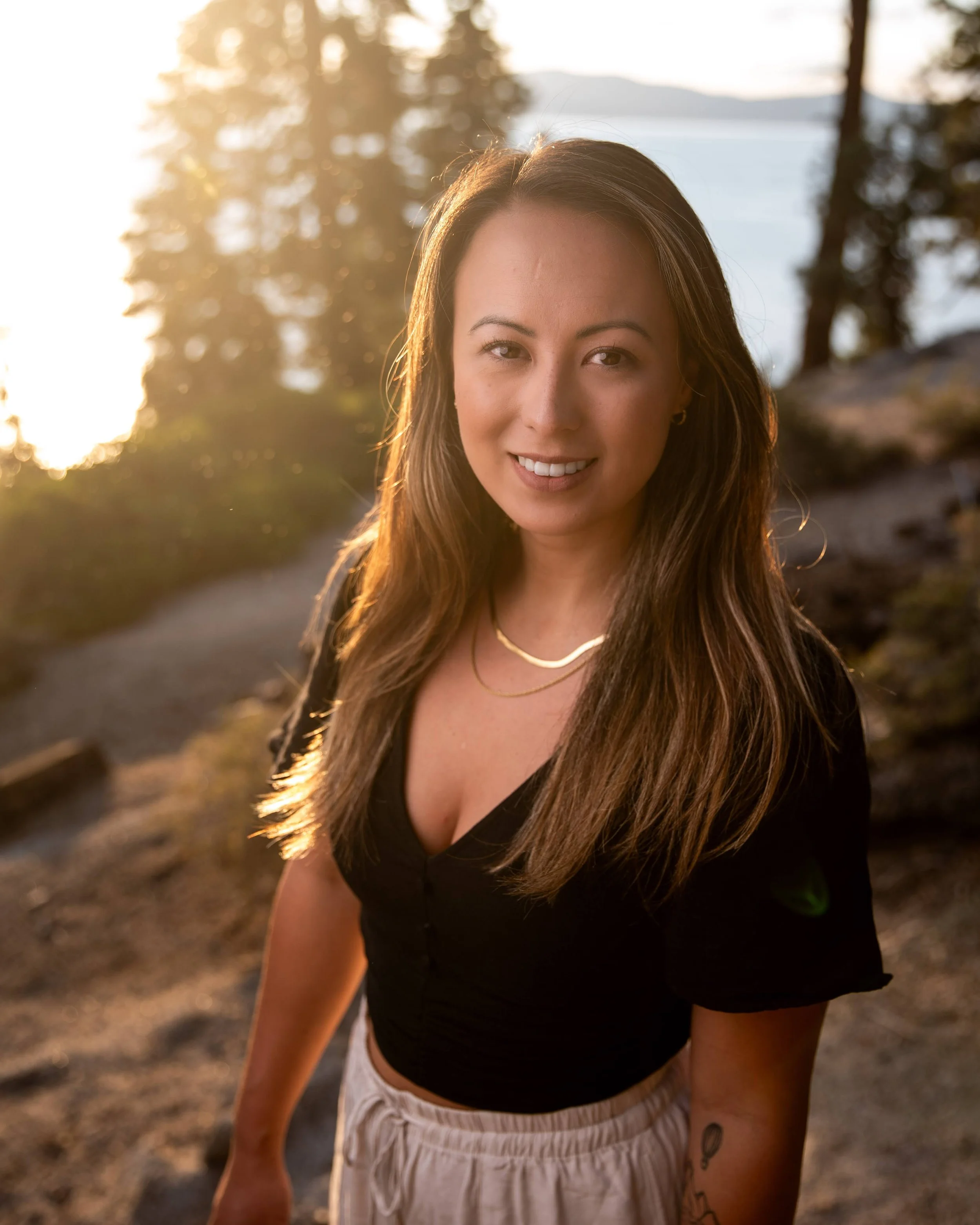 A woman smiling outdoors at sunset, with trees and water in the background.