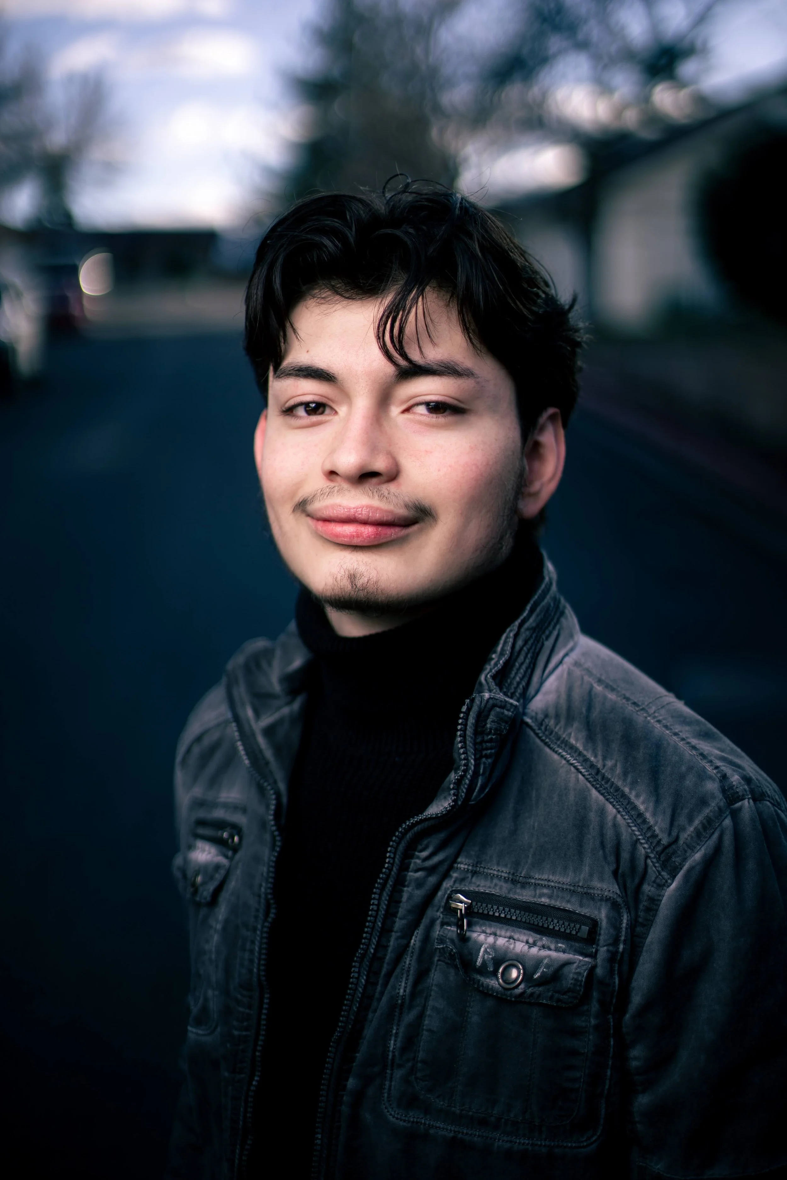A young man with dark hair and light skin, wearing a black turtleneck and a gray jacket, standing outdoors on a darkened street during twilight.